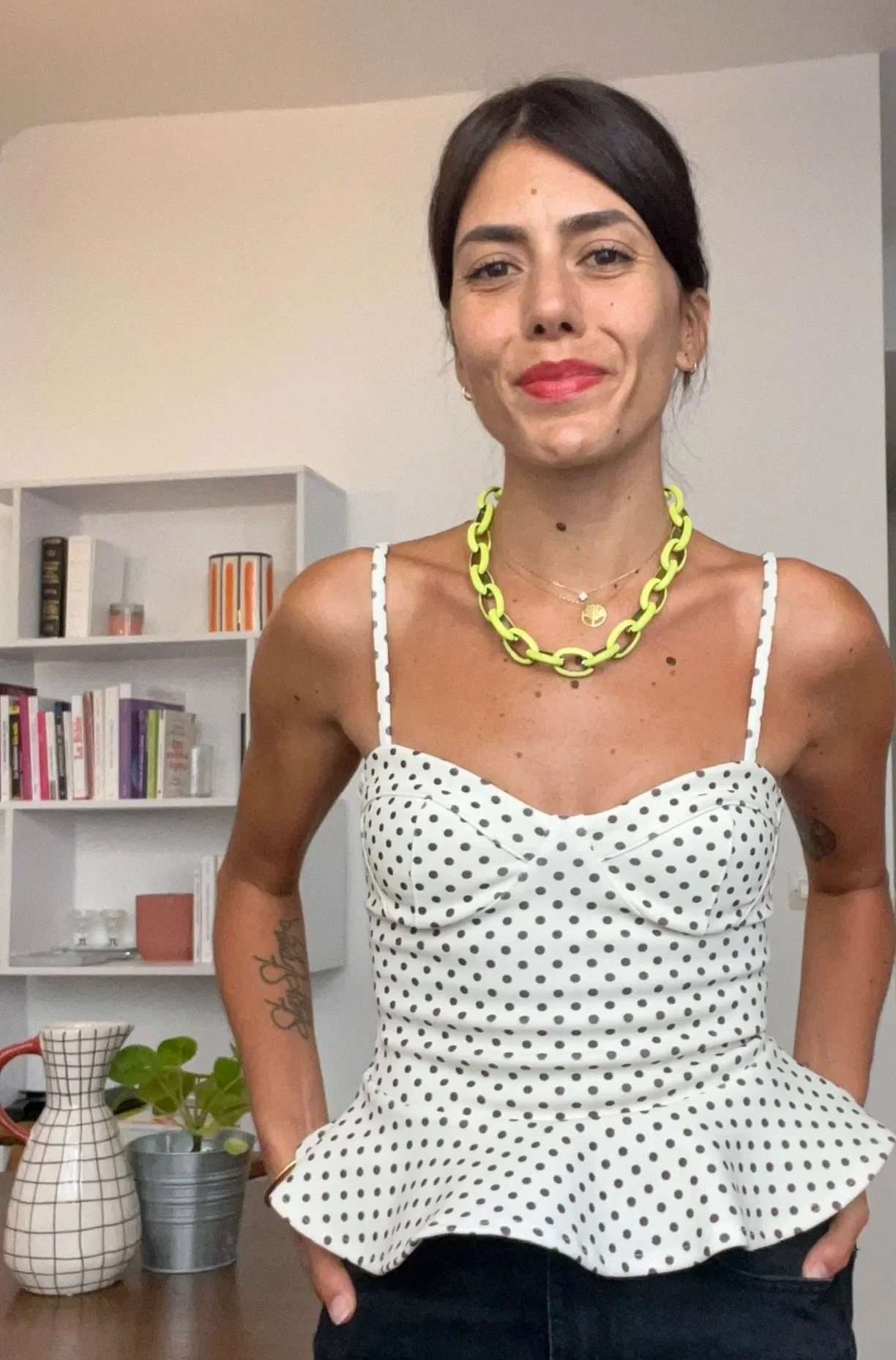 Une photo de Clara, la coach, portant un haut blanc à pois noirs et un collier jaune, posant dans une pièce avec une étagère contenant des livres et des décorations, et des vases sur une table.