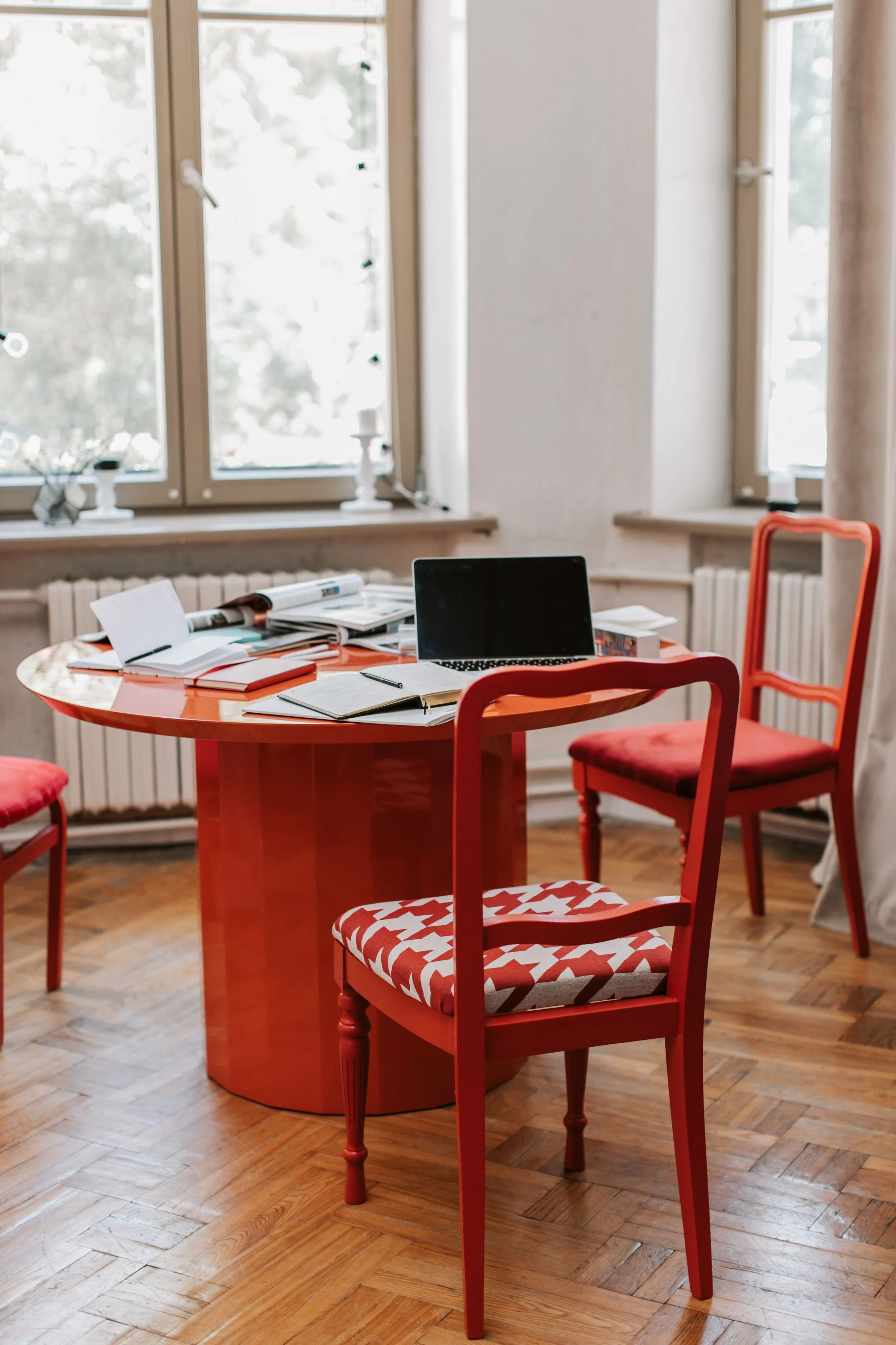 Une salle à manger avec une table ronde orange, entourée de chaises rouges à coussin à motif blanc et rouge, et des fenêtres laissant entrer la lumière naturelle. Rappel code couleur site et couleurs orange pour la vie, le sacré.