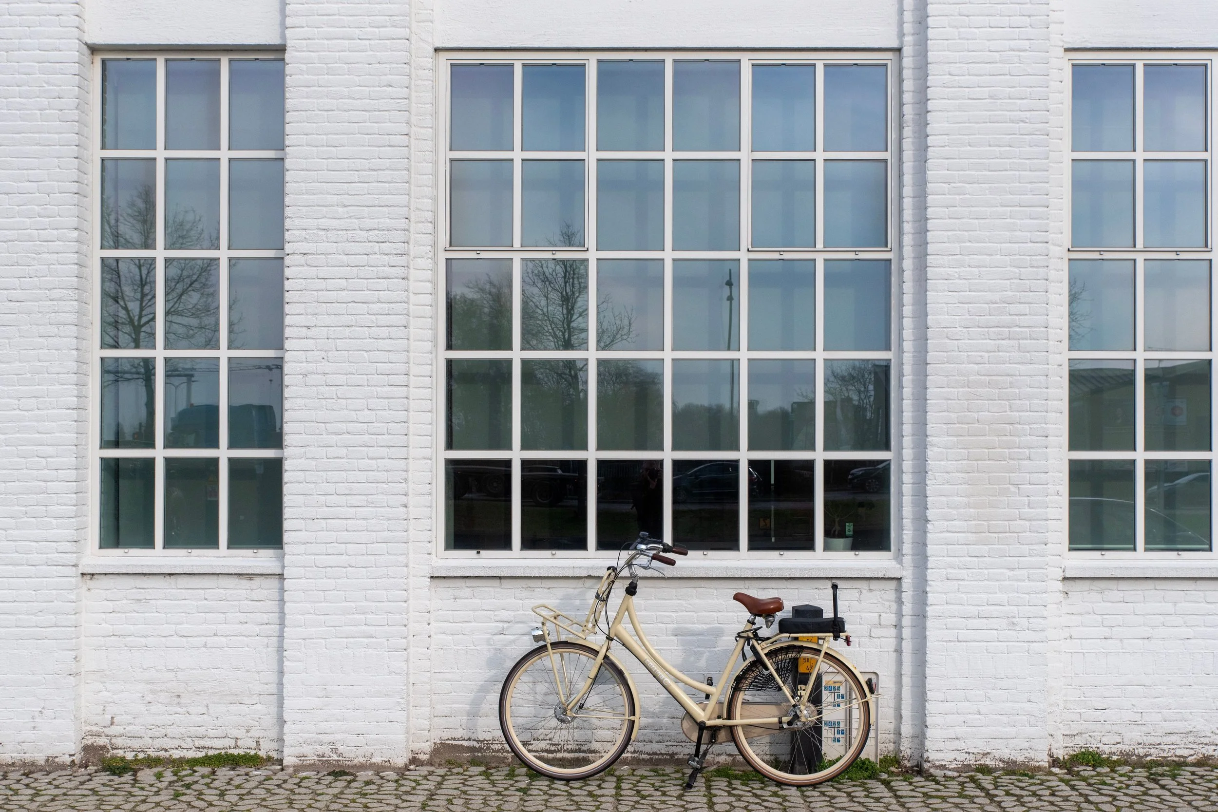 Deux bicycles beige garés devant un mur blanc en briques avec de grandes fenêtres en verre. Le mur a été peint en blanc et la surface au sol est pavée. Cette photo représente le fait de partir à l'aventure de sa propre vie, vers sa mission d'âme.