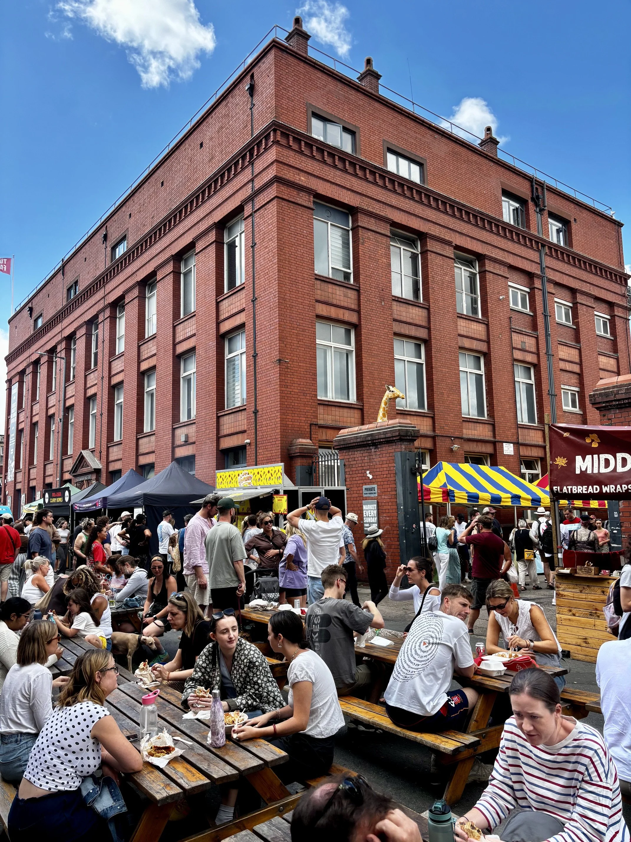 Crowd of people sitting at picnic tables at The Sunday Market with the Tobacco Factory red brick building in the background and food stalls in the foreground.
