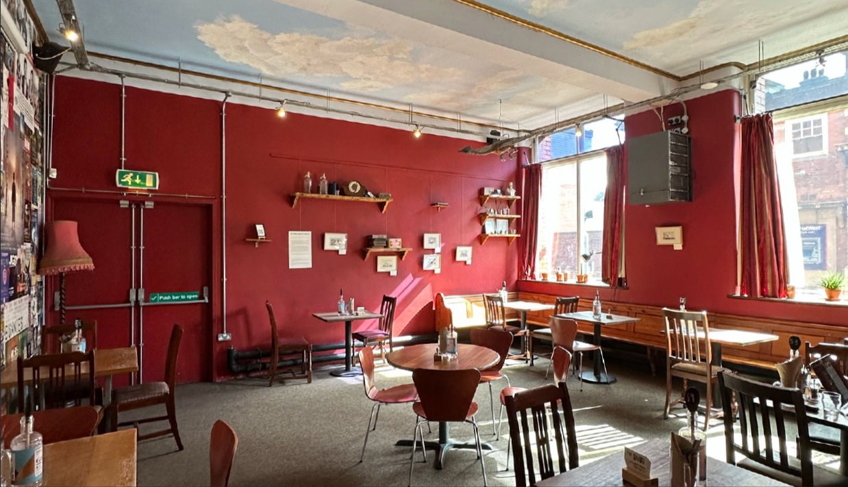 Interior of The Snug showing red walls, wooden shelves, and tables set with condiments. Large windows with red curtains let in natural light. Decor includes framed pictures and a large speaker.
