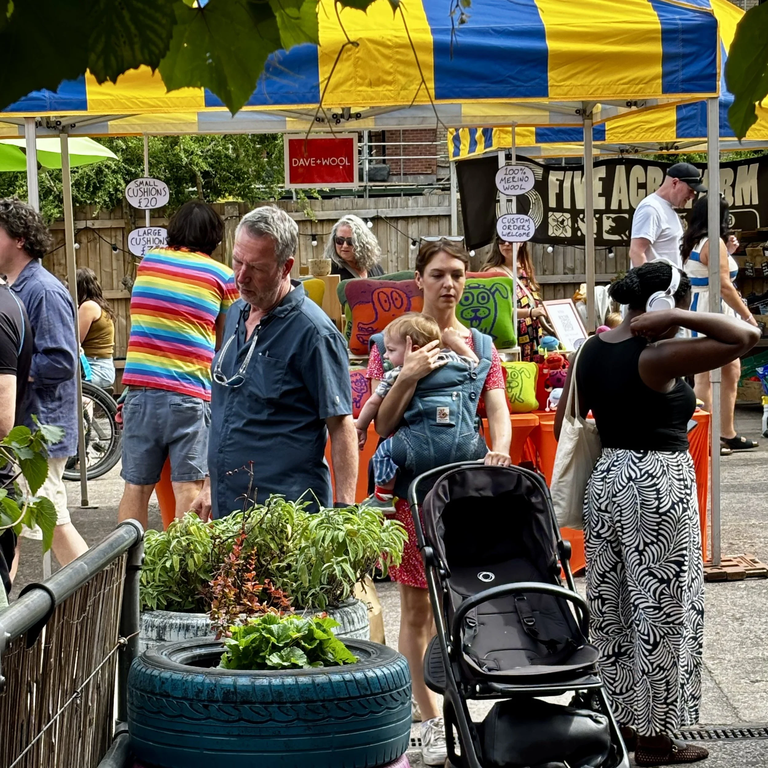 People shopping at the Sunday Market with tents, signs advertising cushions and wool products, and various individuals walking and browsing.