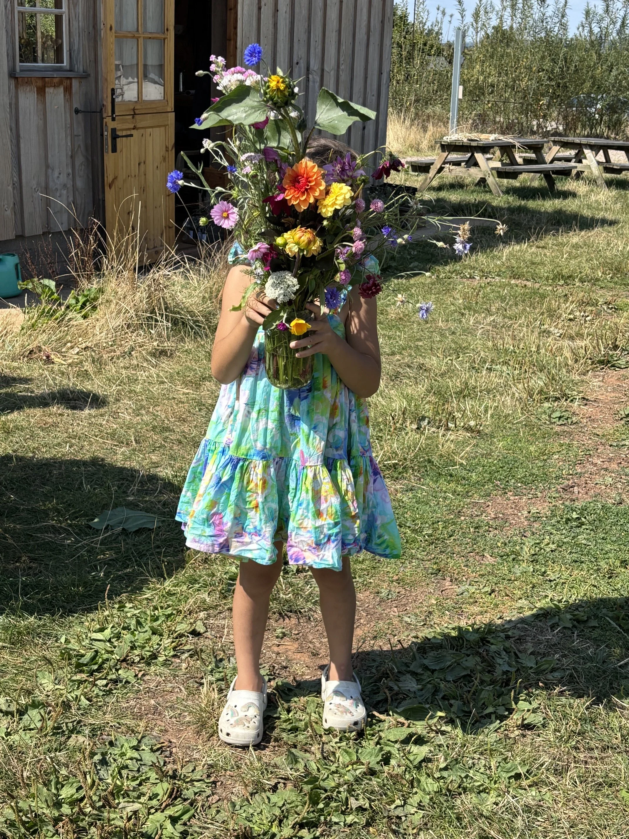 A child holding a colourful bouquet of flowers in front of their face, wearing a tie-dye dress and white shoes, standing on grass in a yard with a wooden shed and picnic tables in the background at 5 Acre Farm
