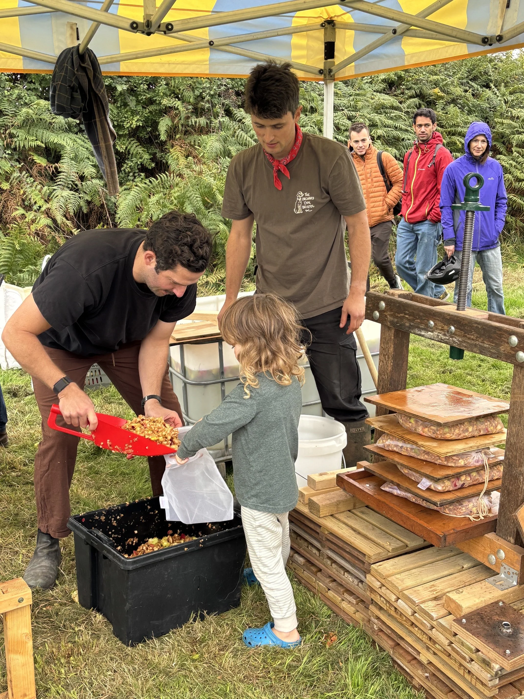 A group of people, gathered at 5 Acre Farm including children and adults, are outdoors under a yellow and white canopy. They are working with what appears to be harvested produce or food, using containers and tools, with some onlookers observing. The