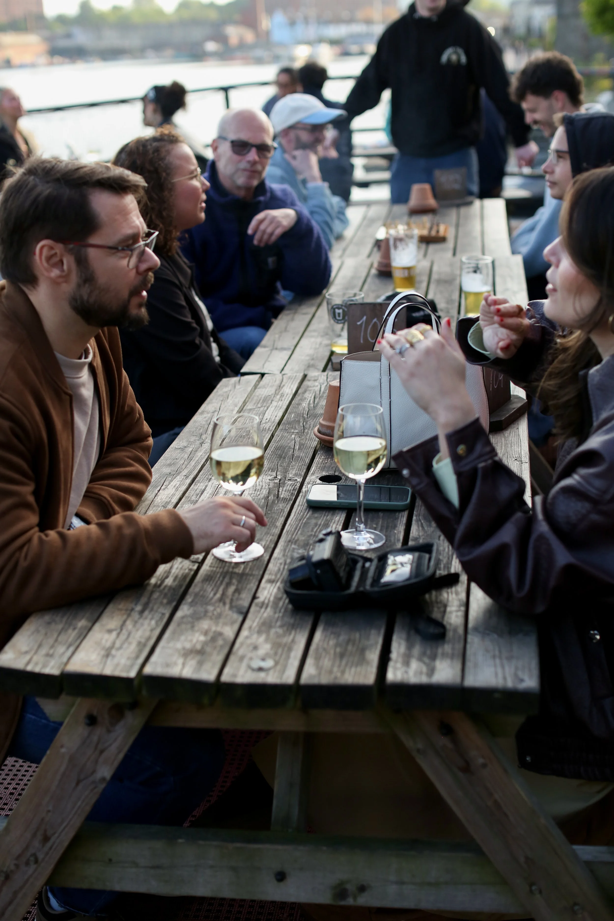 People sitting at a long outdoor wooden table in Grain Barge, enjoying drinks and conversation, with a bridge and trees in the background.