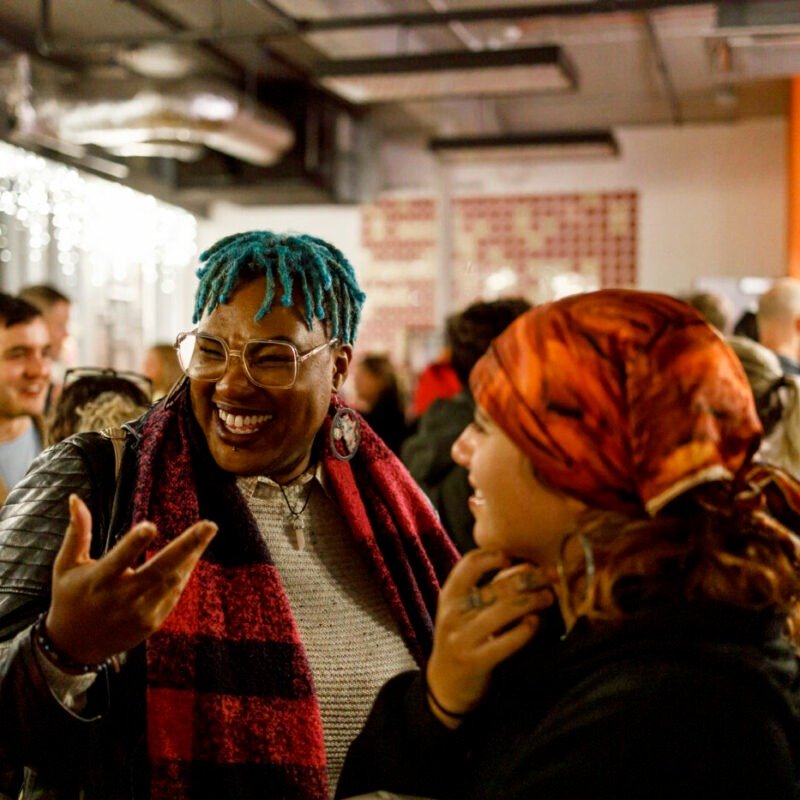 Two women are smiling and talking to each other after an enjoyable performance at Tobacco Factory Theatres. One woman has blue braided hair, glasses, and a red and black scarf. The other woman wears a colourful headscarf.