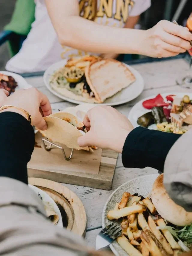People sharing a meal on Grain Barge with dishes including a sandwich, French fries, and various foods on a rustic wooden table.