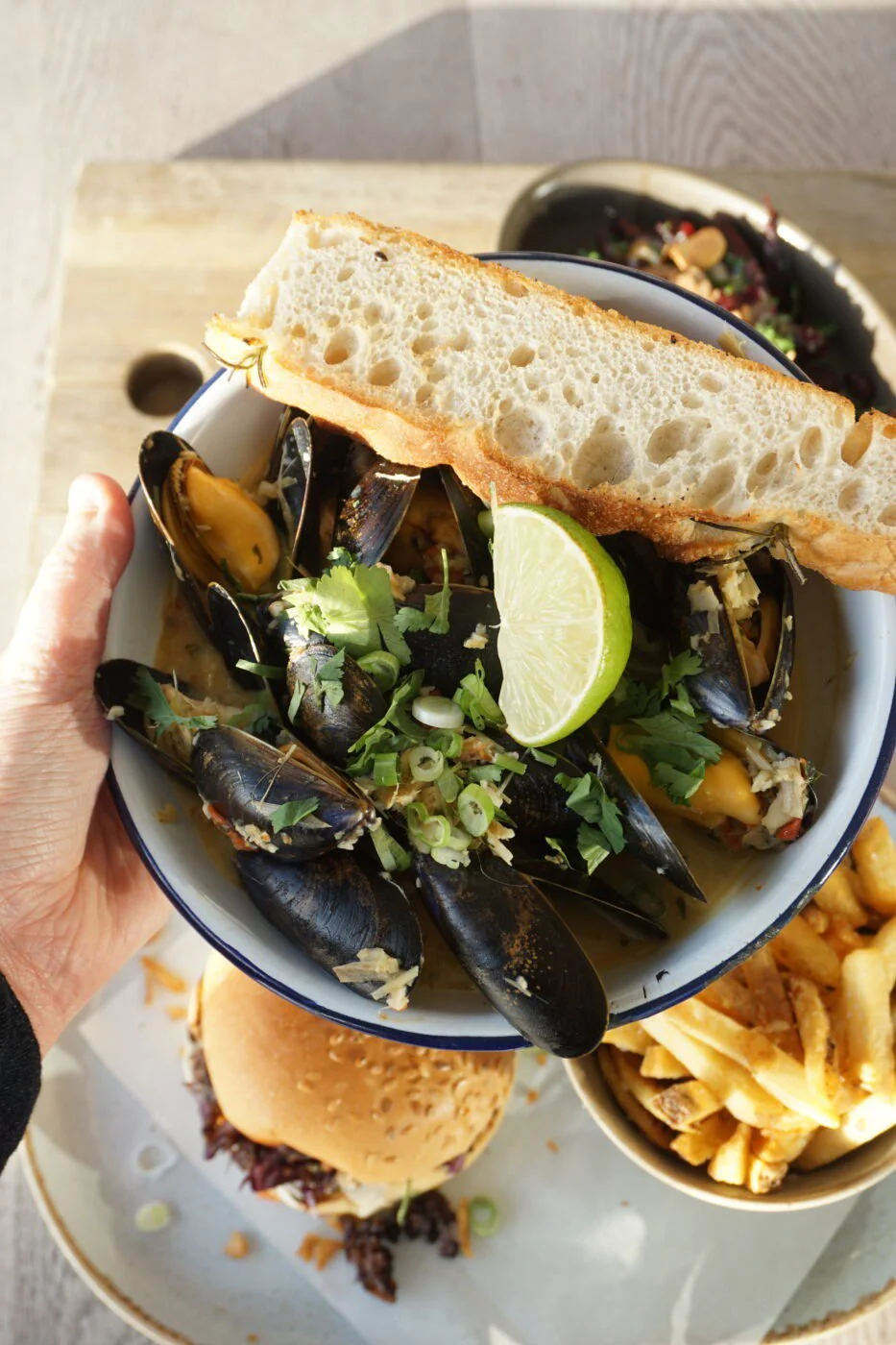 A bowl of steamed mussels garnished with chopped green onions, coriander, and a lime wedge, with slices of bread on top. A hand holds the bowl, with a cheeseburger and fries on a tray below. Grain Barge restaurant moules.