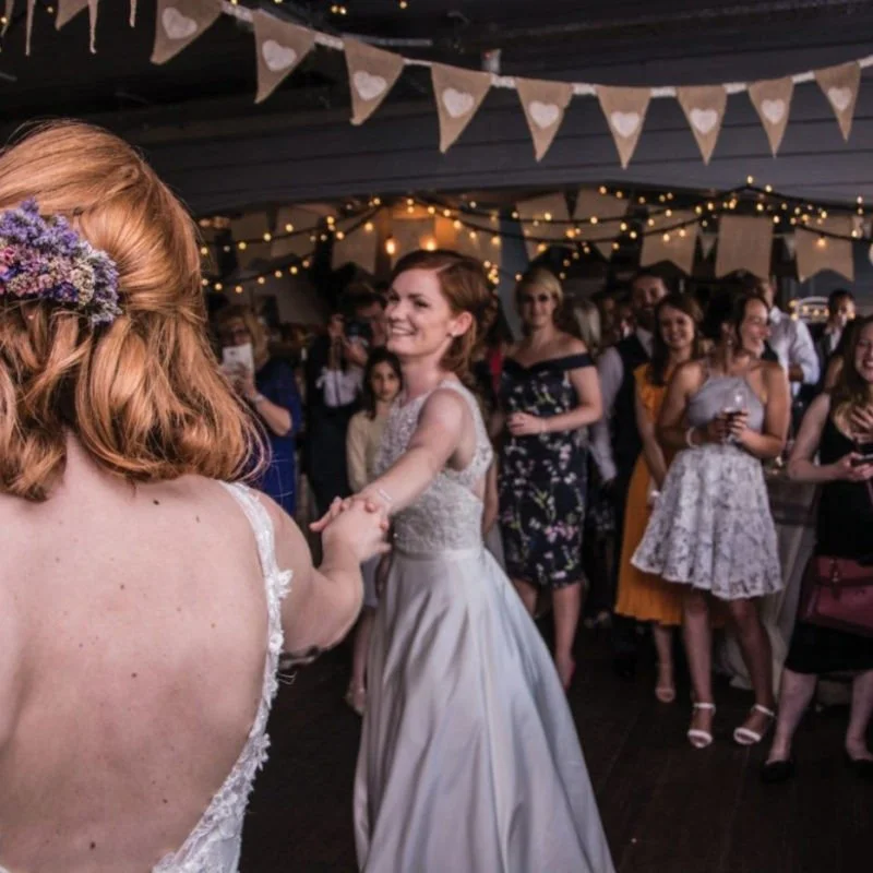 Hire our Space. A wedding reception  in the Hold Bar on Grain Barge, with a bride dancing with a guest, surrounded by guests under decorated banners and string lights.