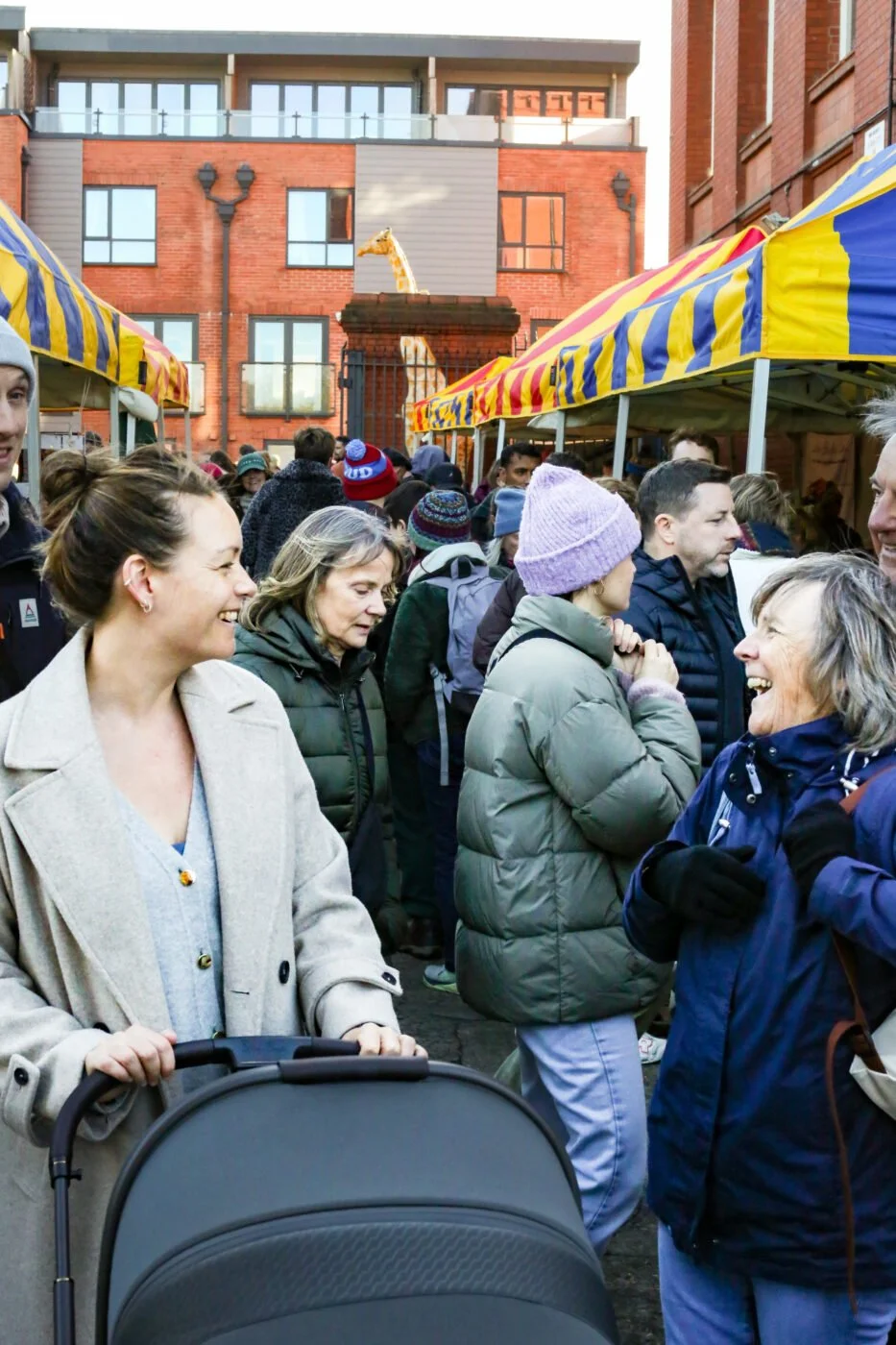 People smiling and chatting at the Sunday Market with yellow and blue tents, with the red brick Tobacco Factory building and a giraffe statue in the background.