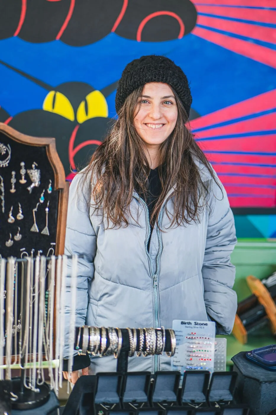 One of the Sunday Market stallholders. Young woman with long hair, wearing a black knit hat and light grey jacket, standing behind a jewellery display.