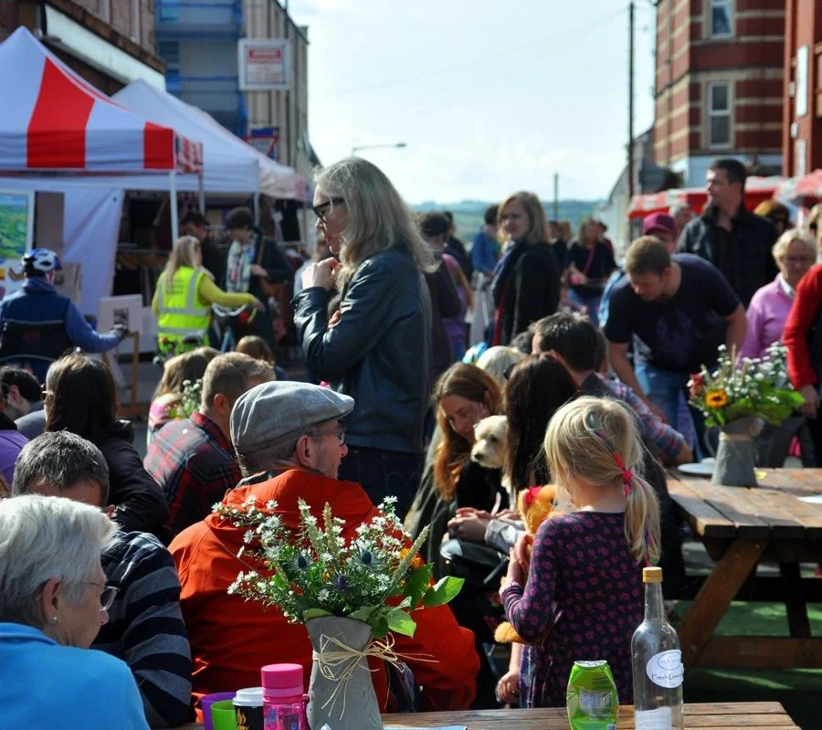 Crowd of people gathered outdoors at the Sunday Market with tents, tables, flowers, and a dog watching in the background.