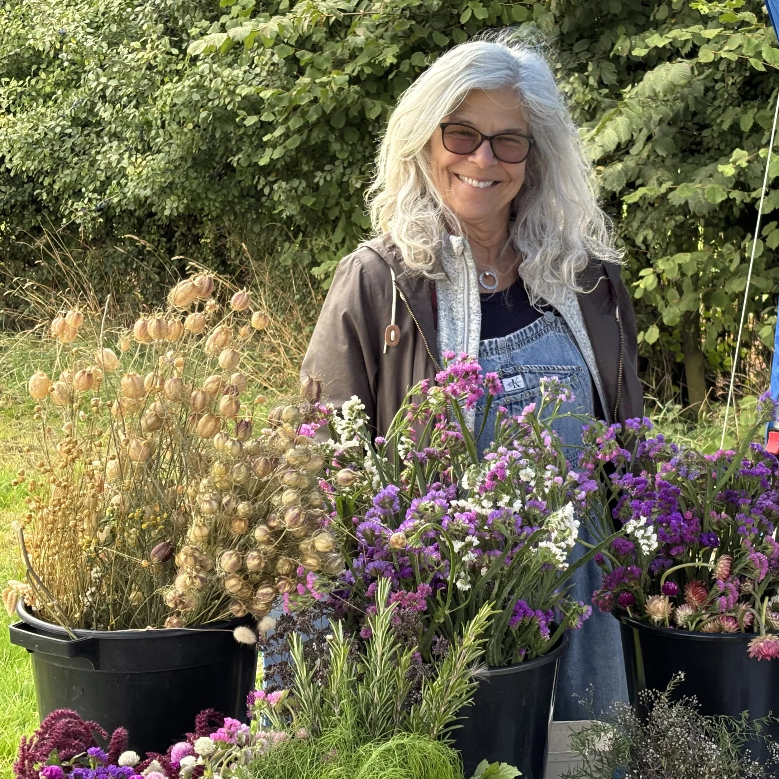 Multiple containers filled with various colourful flowers outside at 5 Acre Farm with green bushes in the background.