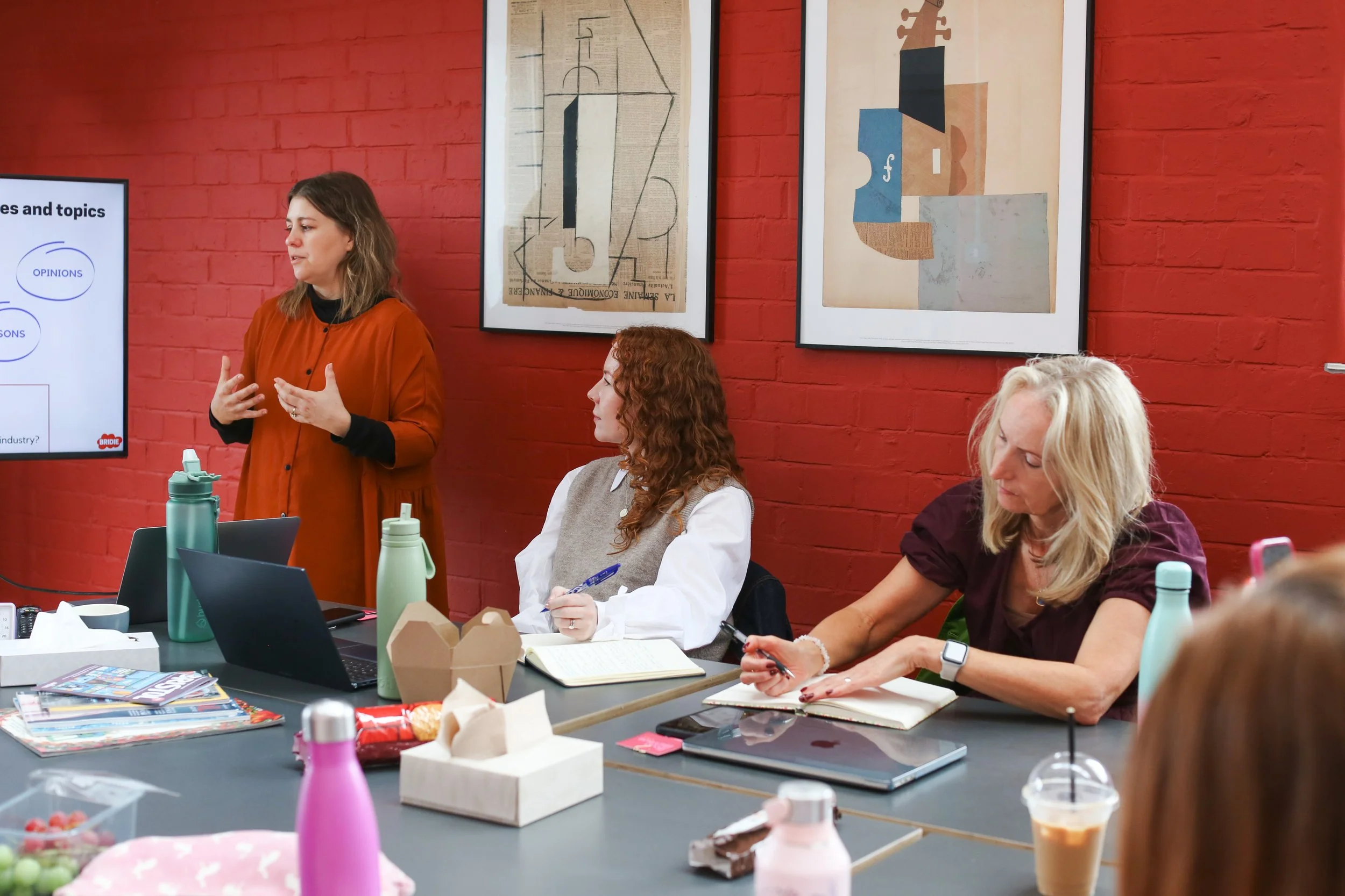 A woman in an orange shirt is standing and speaking to a group of people in a meeting room with a red brick wall. Three women are seated at the table, one with red hair taking notes, another with blonde hair looking at her phone, and a third with long hair wearing a white shirt. The table has laptops, notebooks, drinks, and snacks. Art posters hang on the brick wall.