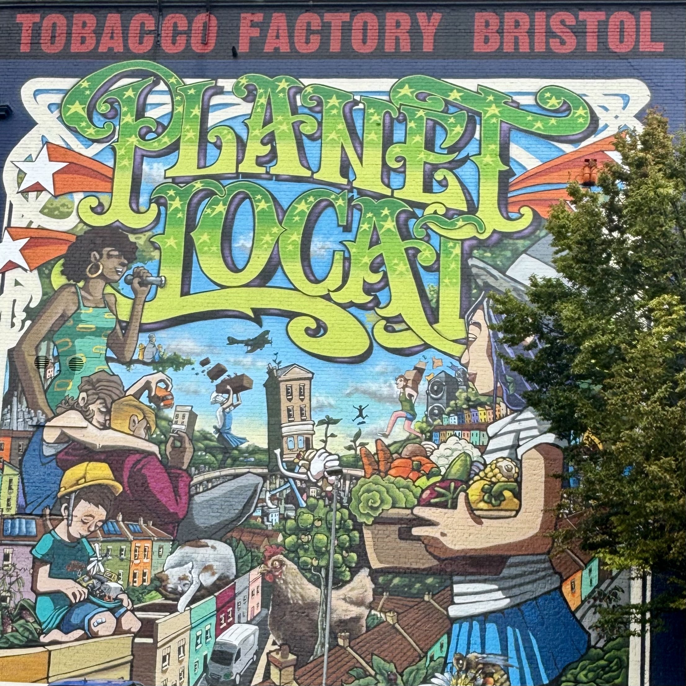 A colourful mural on a brick wall featuring the words 'Planet Local' in green and yellow lettering surrounded by illustrations of diverse people, buildings, trees, and flags, with a sign above reading 'Tobacco Factory Bristol'.