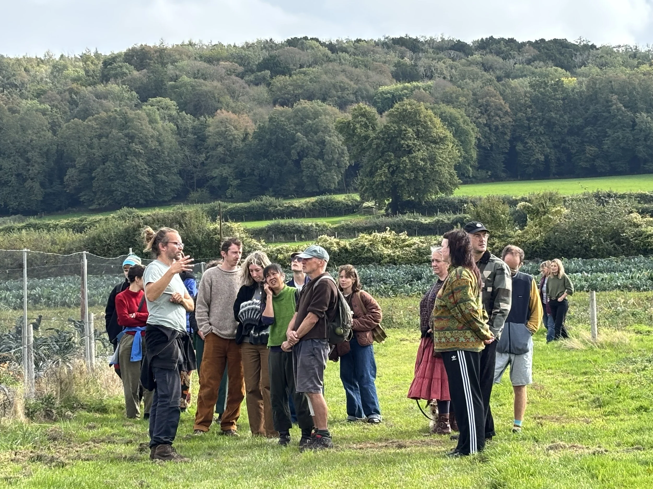 A group of people gathered outdoors at 5 Acre Farm in front of a lush green hillside, engaging in conversation.