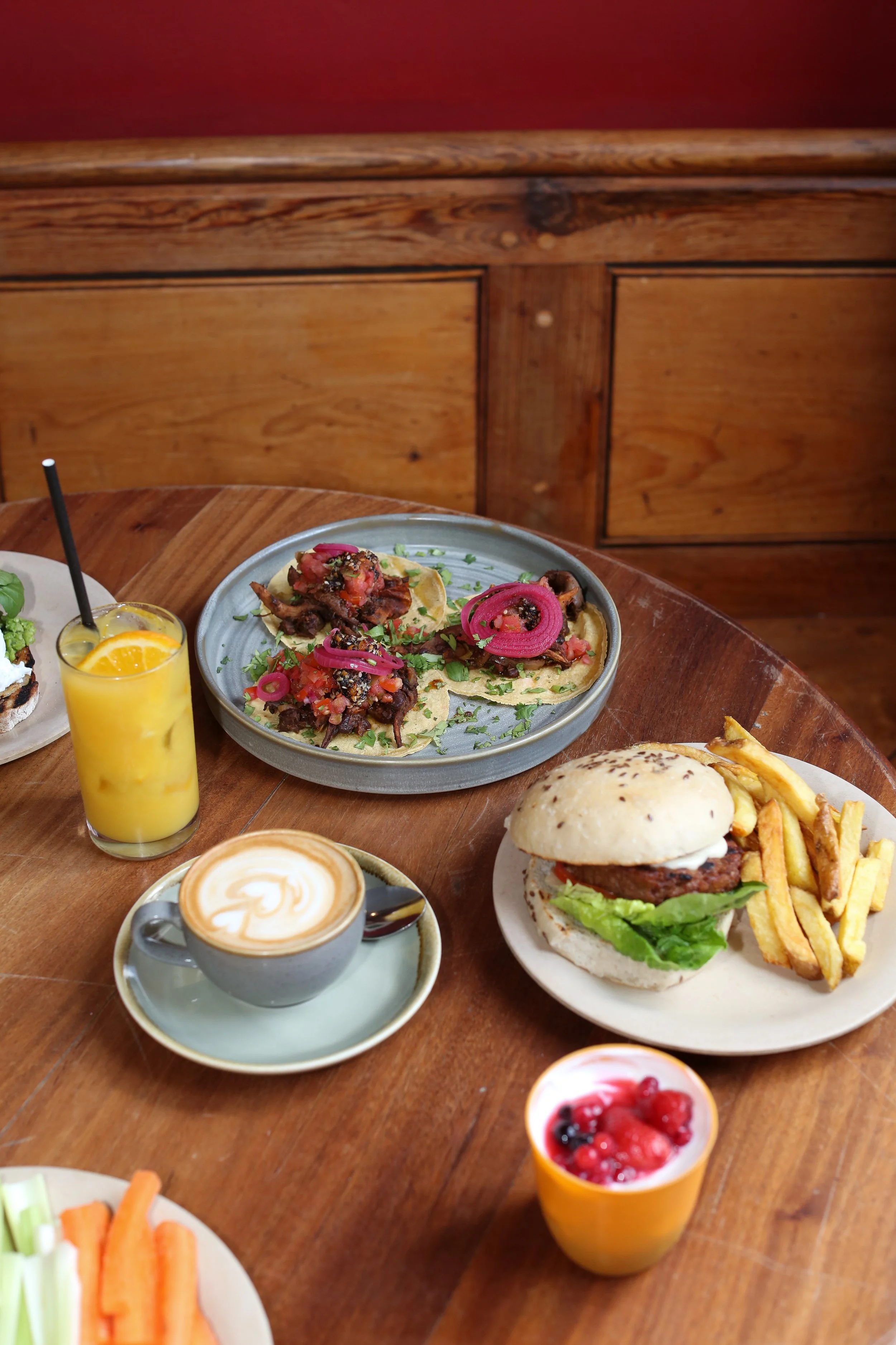 A plate of tacos with meat, onions, and herbs, a cheeseburger with fries, a cappuccino with latte art, a glass of orange juice with a straw, and bowls of fruit and vegetables on a wooden table with a wooden wall in the background.