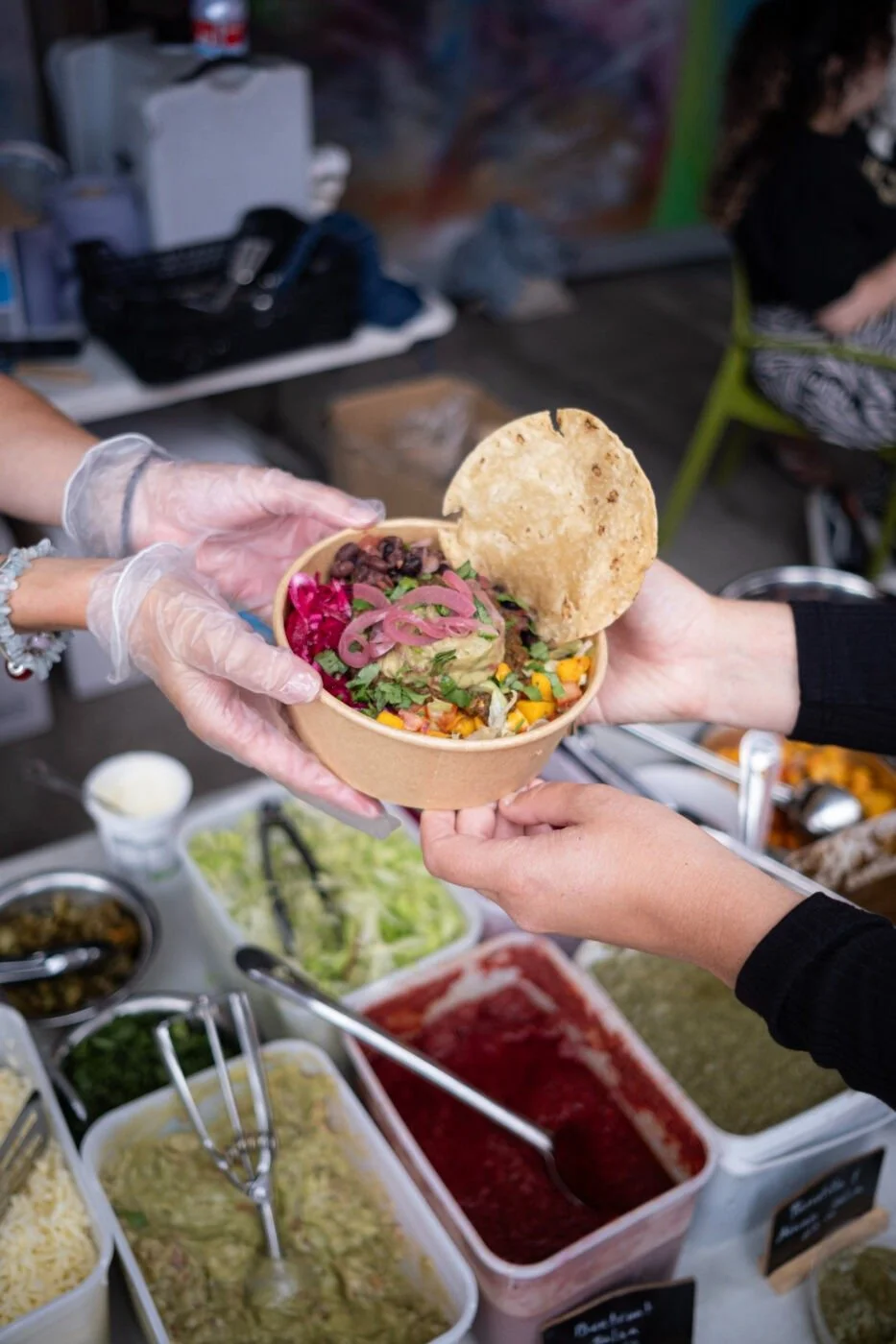 Person handing a bowl of salad with toppings and a large tortilla chip to another person at a food stand, with various containers of ingredients in the background.