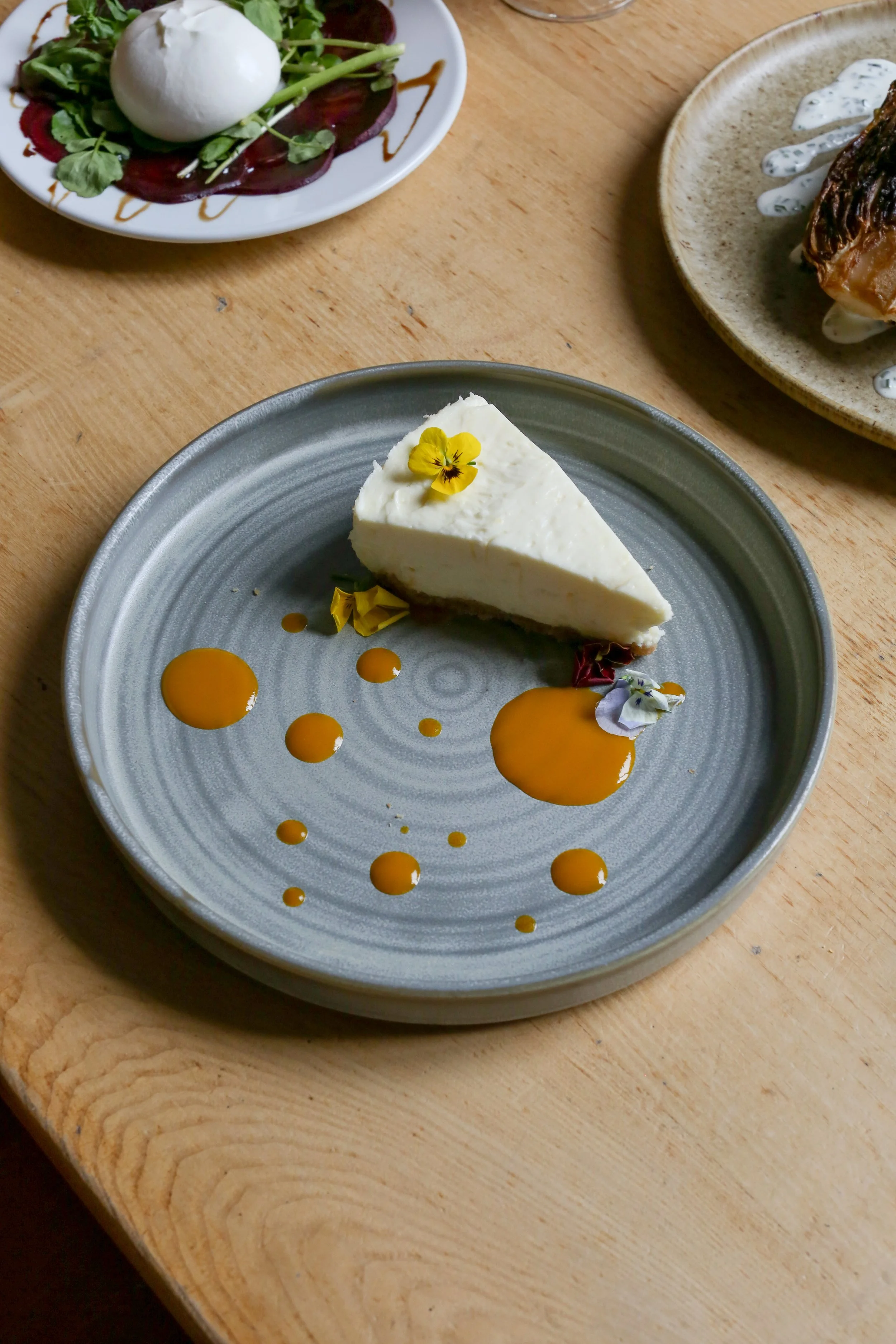 A slice of cheesecake garnished with edible flowers and drizzled with caramel sauce, served on a gray plate. Partially visible plates with salad and meat are in the background.