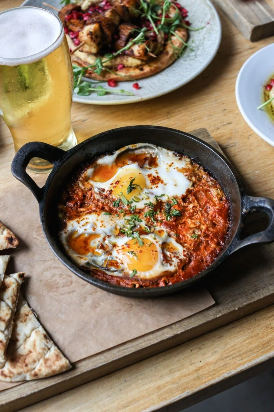 A skillet with shakshuka featuring eggs in tomato sauce, garnished with herbs, on a wooden table. Nearby is a glass of beer, a plate of grilled meat skewers with greens, and various plates of food.