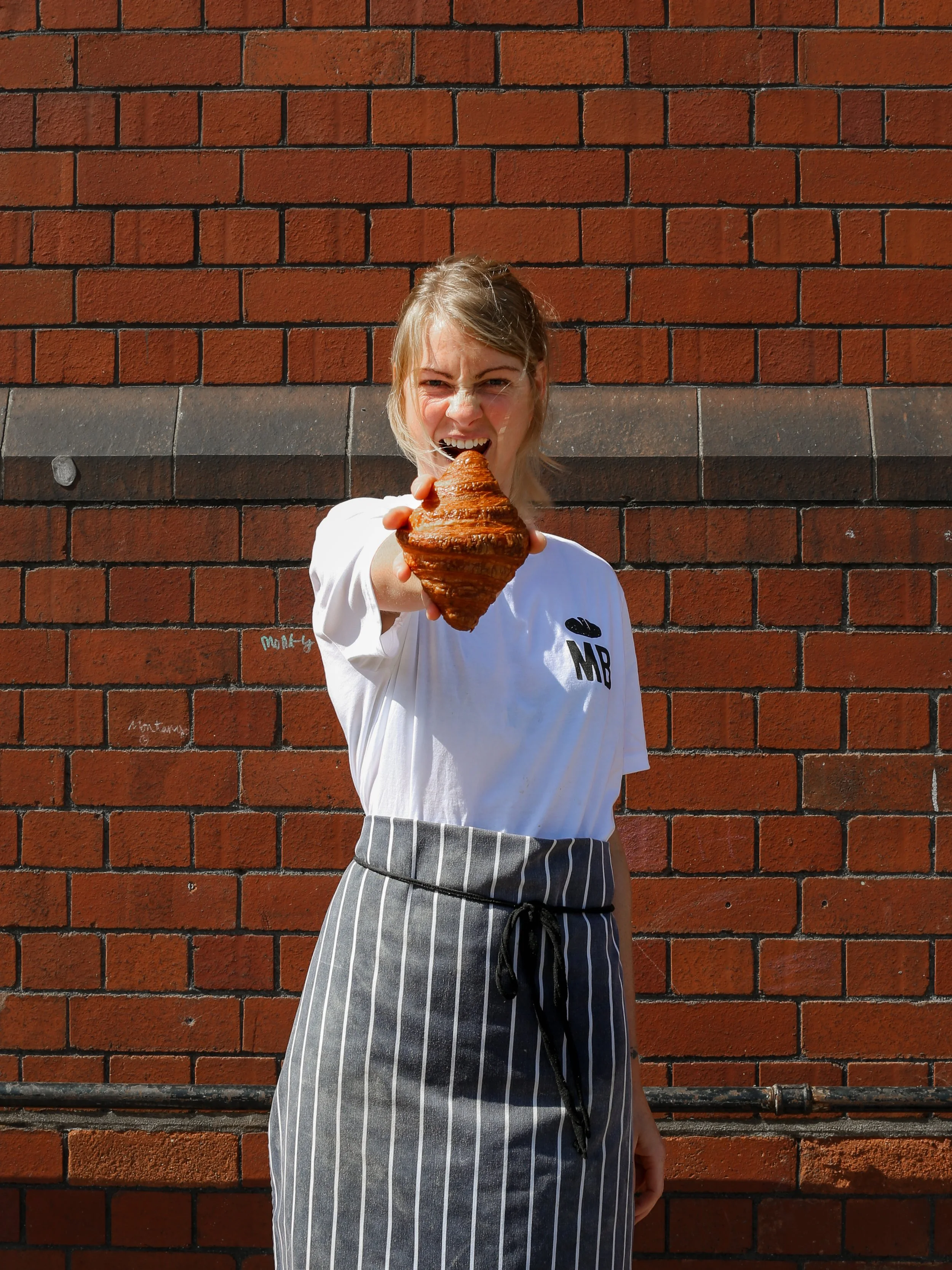 A woman standing in front of the Tobacco Factory and Mark's Bakery red brick wall, holding a croissant and pretending to bite it, wearing a white t-shirt with Mark's Bakery logo on and striped apron.