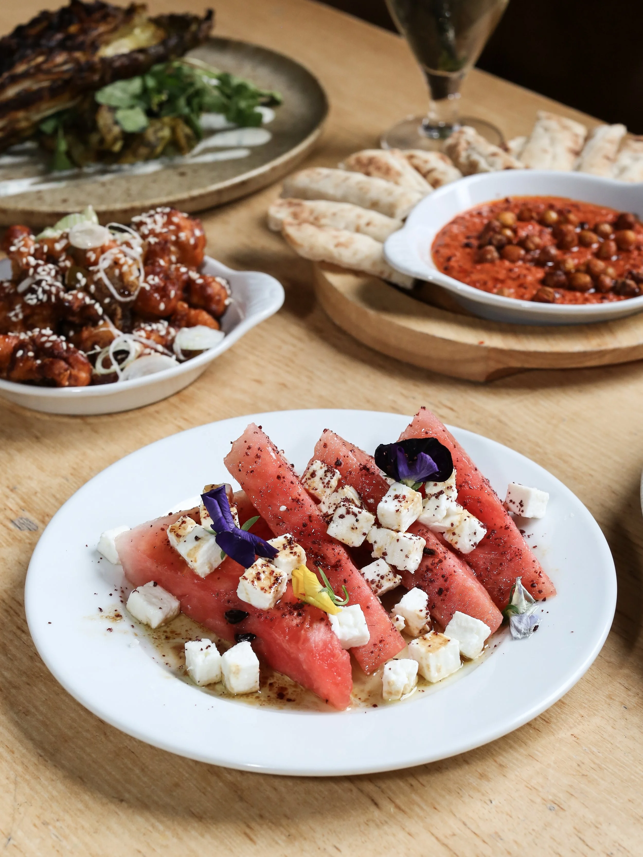 Close-up of a white plate with sliced watermelon, feta cheese, and edible flowers on top, on a wooden table.
