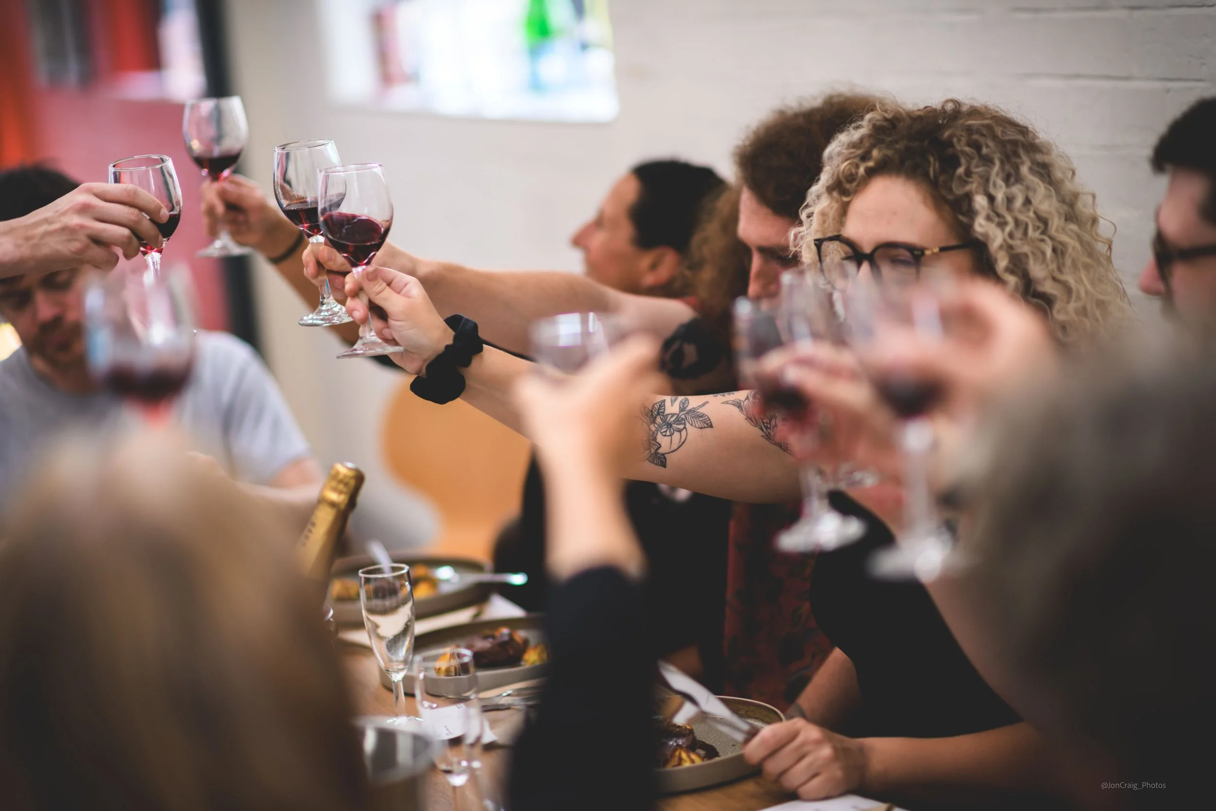 People who have hired The Loft space for a private event, sitting around a table enjoying a toast with glasses of red wine at a social gathering or celebration.