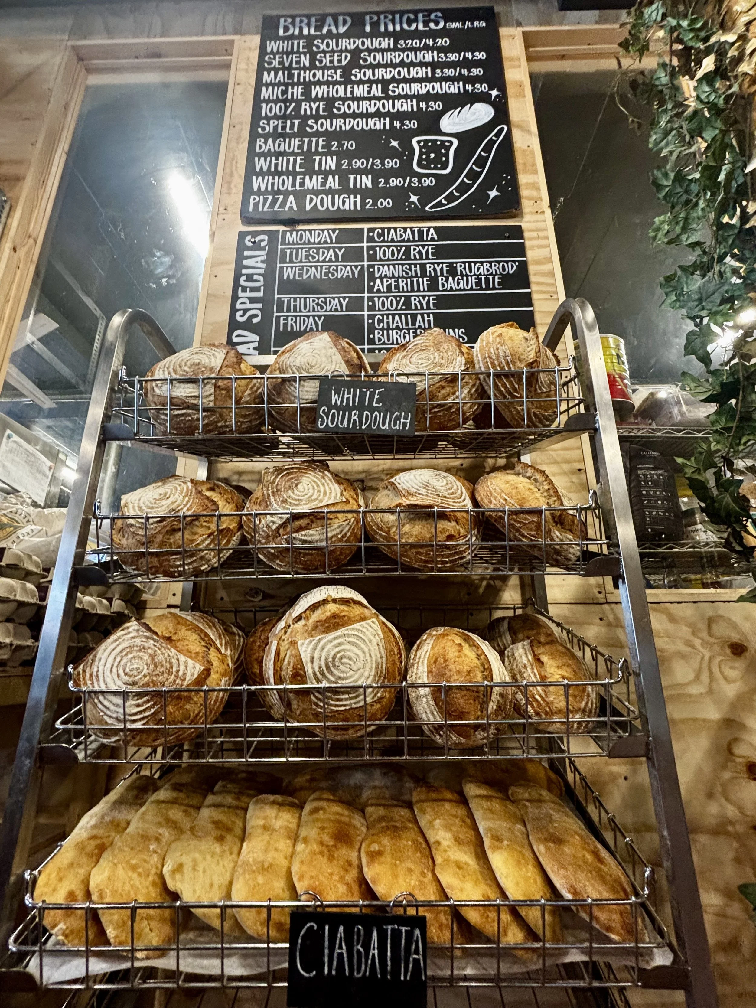 Display of various types of Mark's Bakery loaves and bread batons on a metal rack with a handwritten sign labeled "Ciabatta" at the front. Behind the rack, there are two black menus on a wooden wall that list bread prices and weekly specials includin