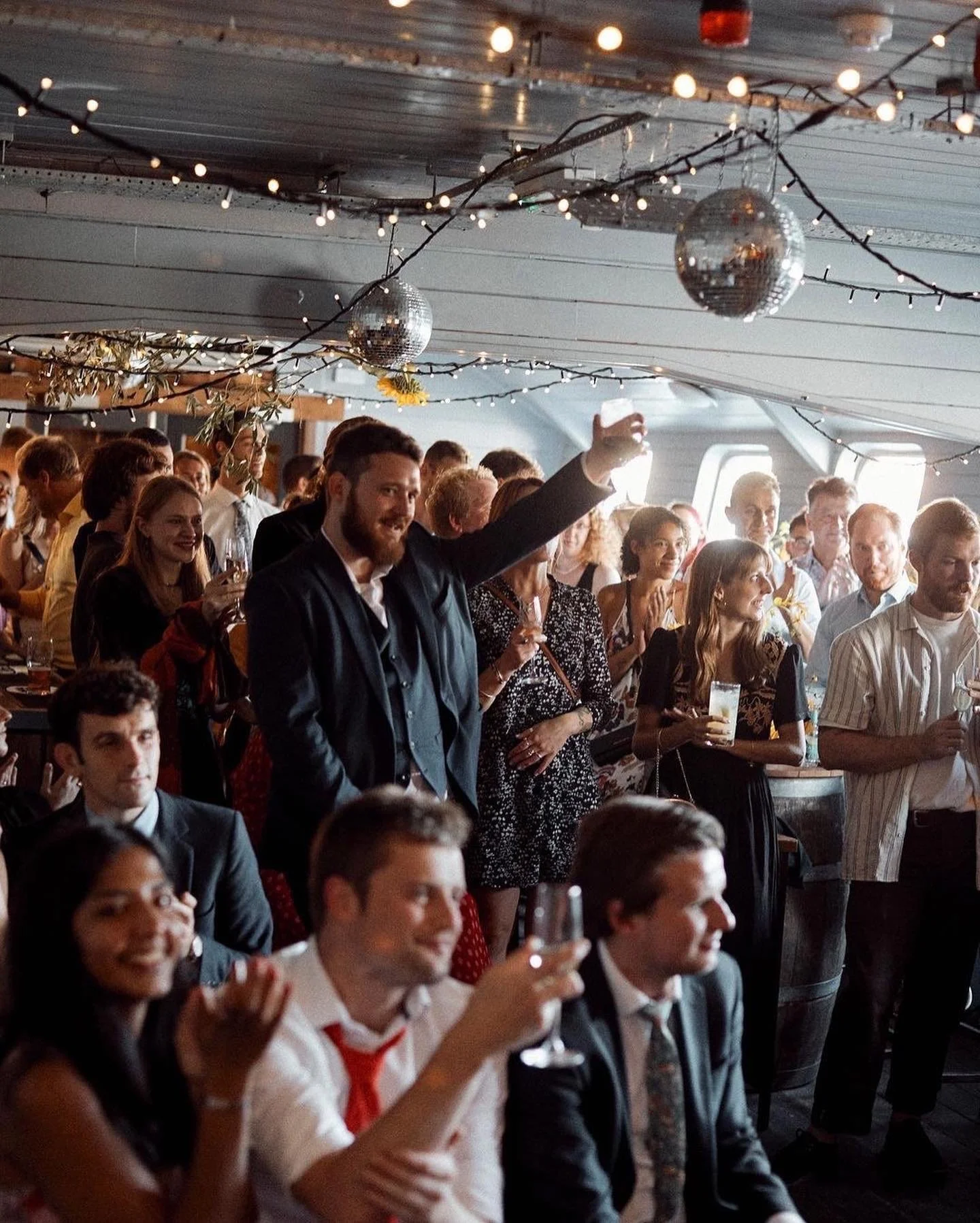 People who have hired thepartying on The Grain Barge. A man with a beard and dark jacket raising a glass in a toast at a social gathering in a decorated venue with fairy lights and disco balls, surrounded by smiling and clapping people holding drinks