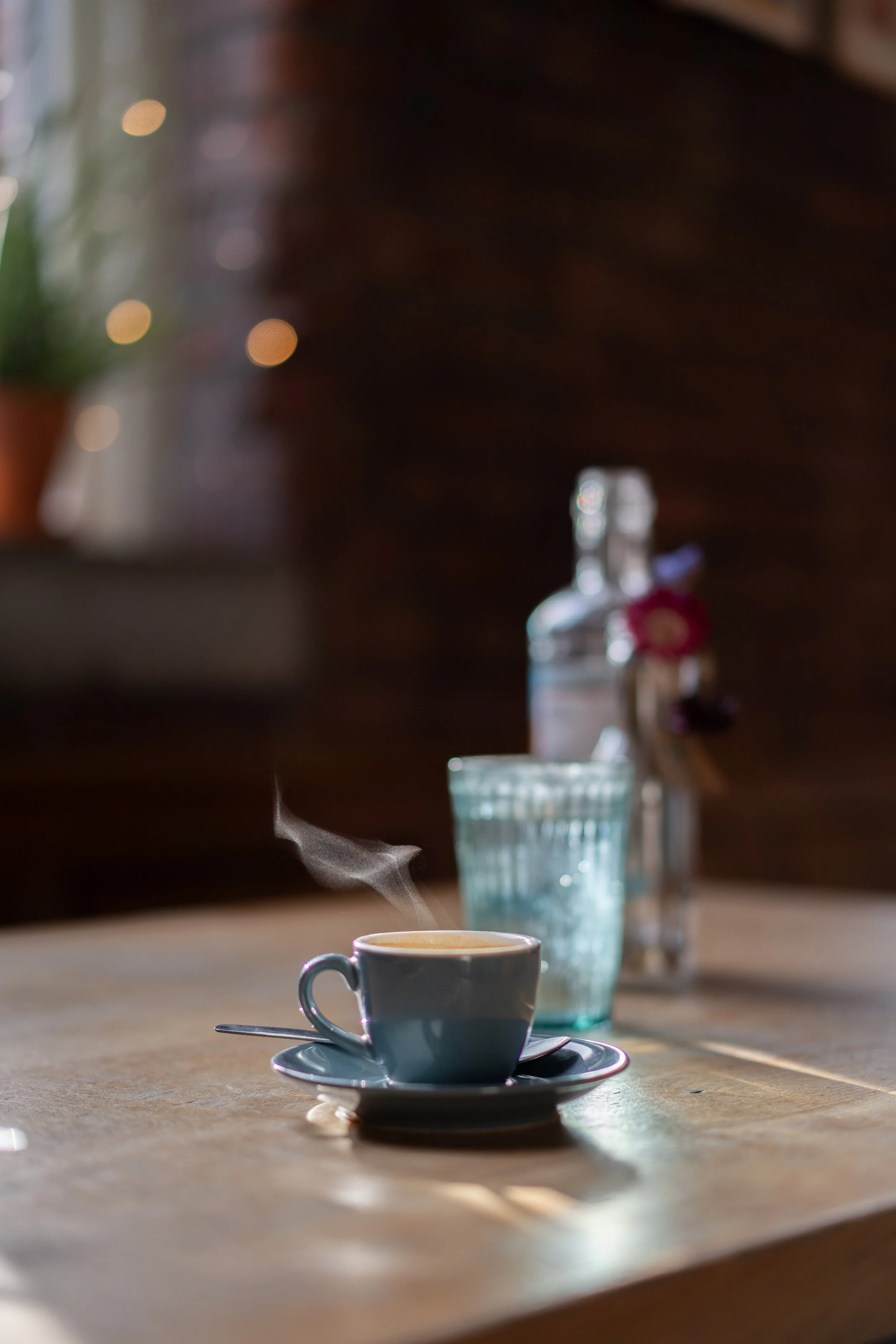 A steaming cup of coffee on a saucer with a spoon, placed on a wooden table. In the background, a glass of water and a salt shaker are visible, with soft lighting creating a cozy atmosphere in The Snug