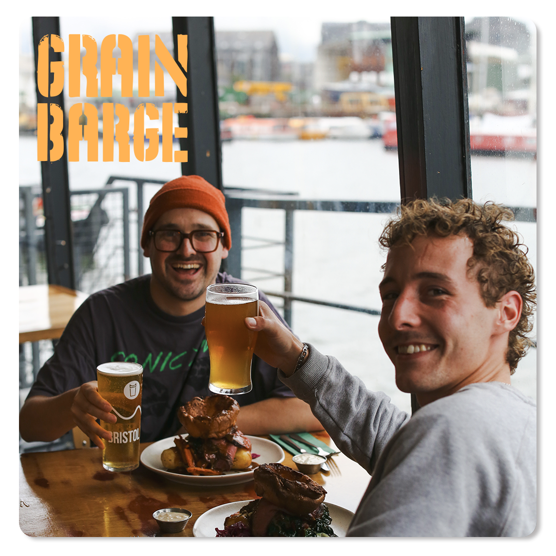 Two smiling men enjoying beers and food at a restaurant with large windows overlooking a body of water and boats from inside the Grain Barge restaurant and bar. Available for private hire is the Hold Bar