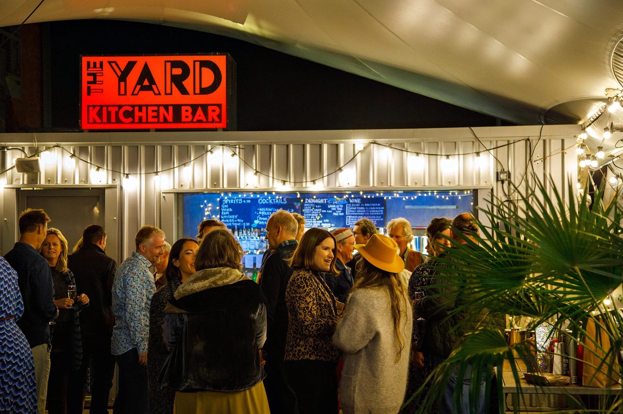 People gathered at an outdoor bar under string lights at night, with a bright sign that reads 'The Yard Kitchen Bar'