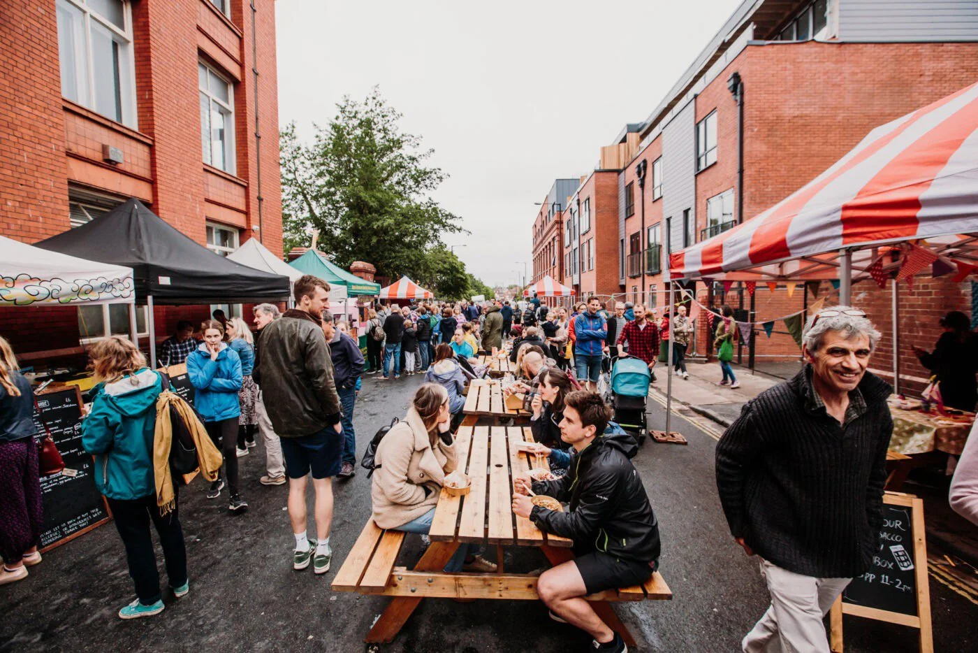 Crowd of people at the Sunday Market with food stalls and tables, red brick buildings on both sides, cloudy sky.