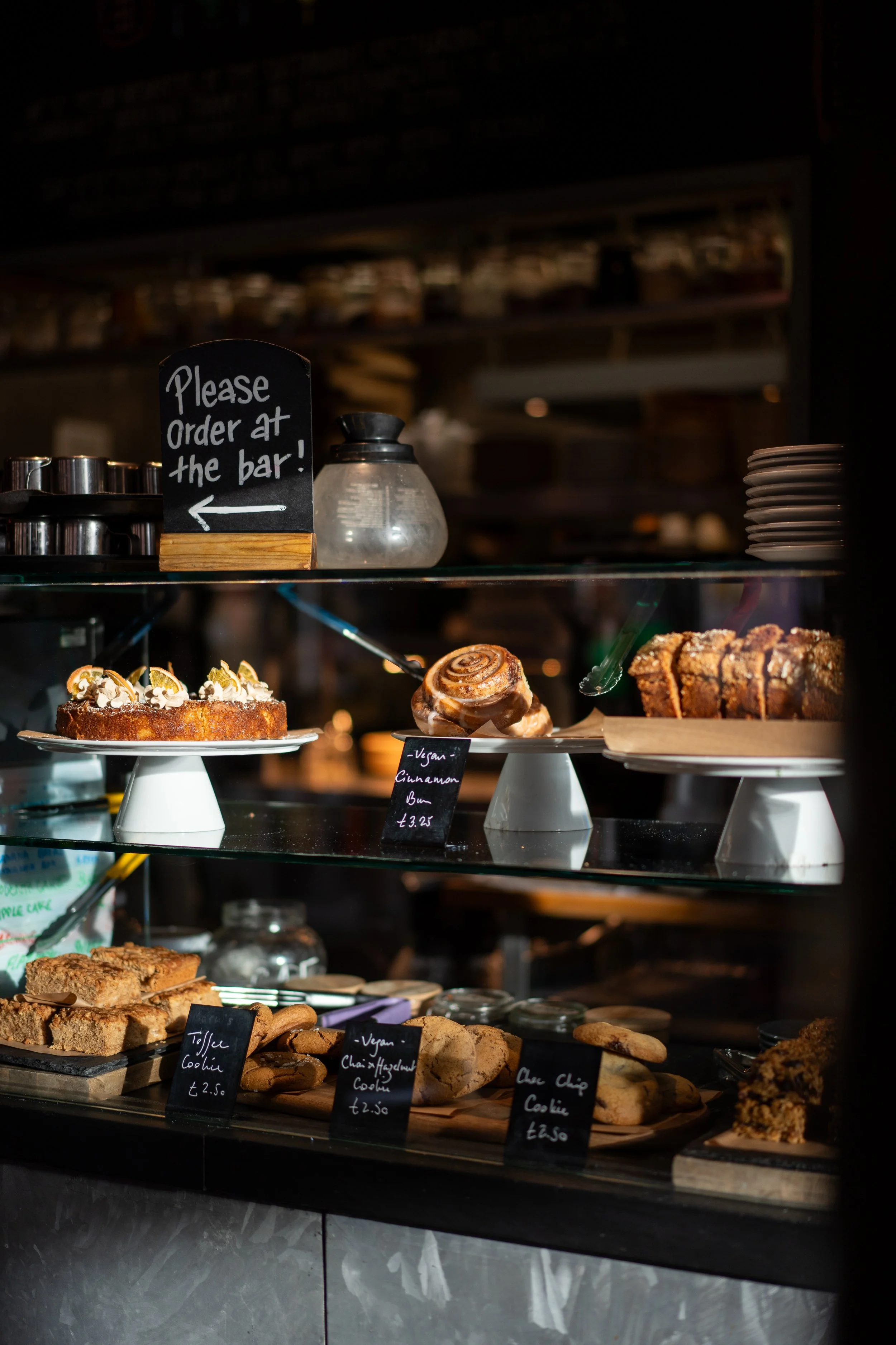 Cafe Bar counter display case with various desserts including cookies and cakes, with small black chalkboard signs labeling each item and a blackboard sign instructing to order at the bar.