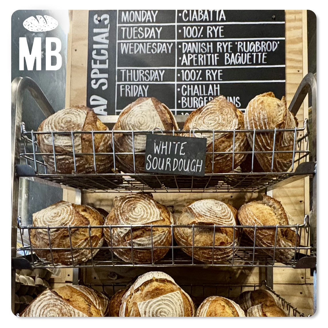 Mark's Bakery - Shelf with multiple loaves of white sourdough bread on metal racks, and a chalkboard sign in the background listing daily specials.