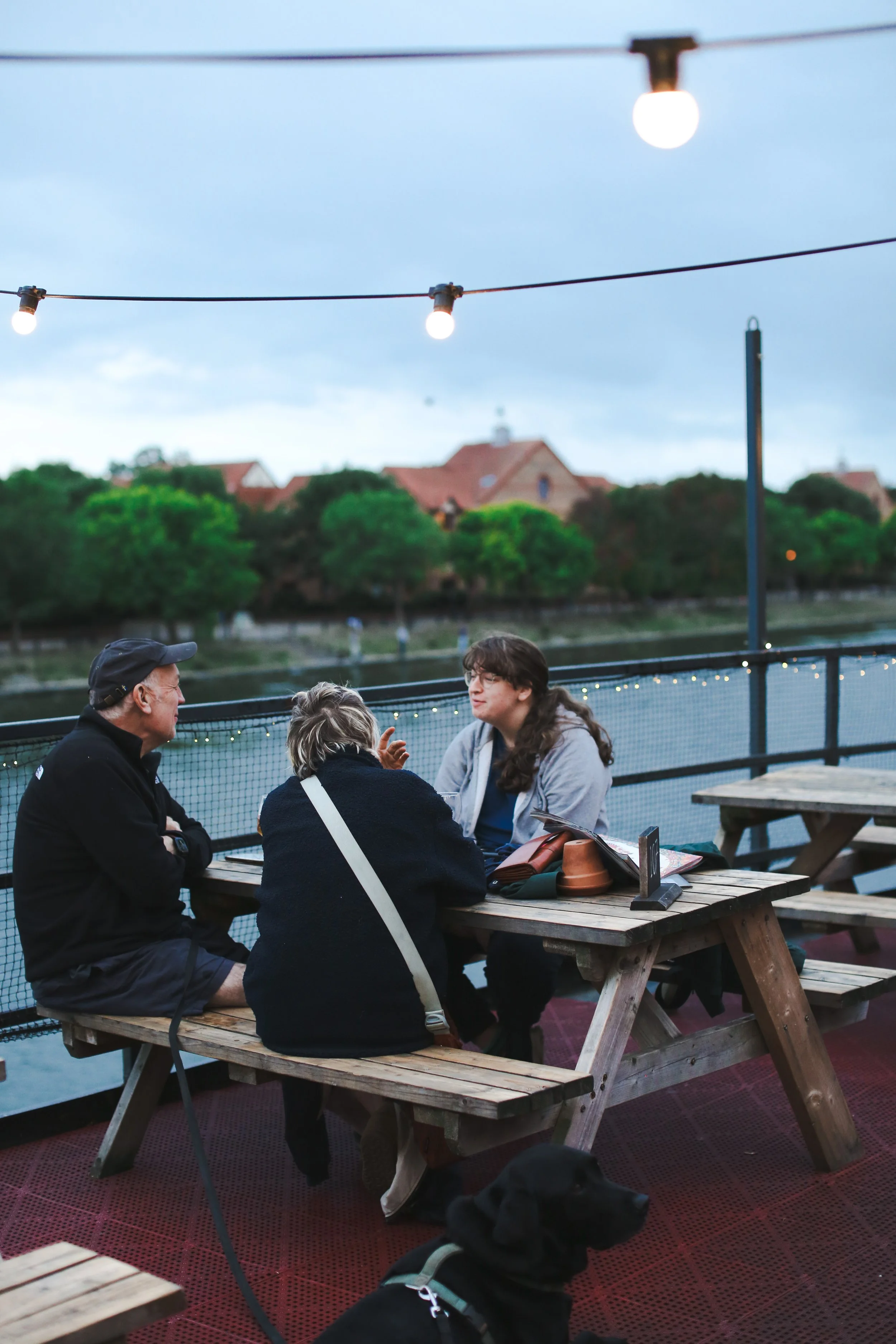 People sitting outdoors at Grain Barge's wooden picnic tables, engaged in conversation, under string lights with the harbour and houses with red roofs in the background during dusk.