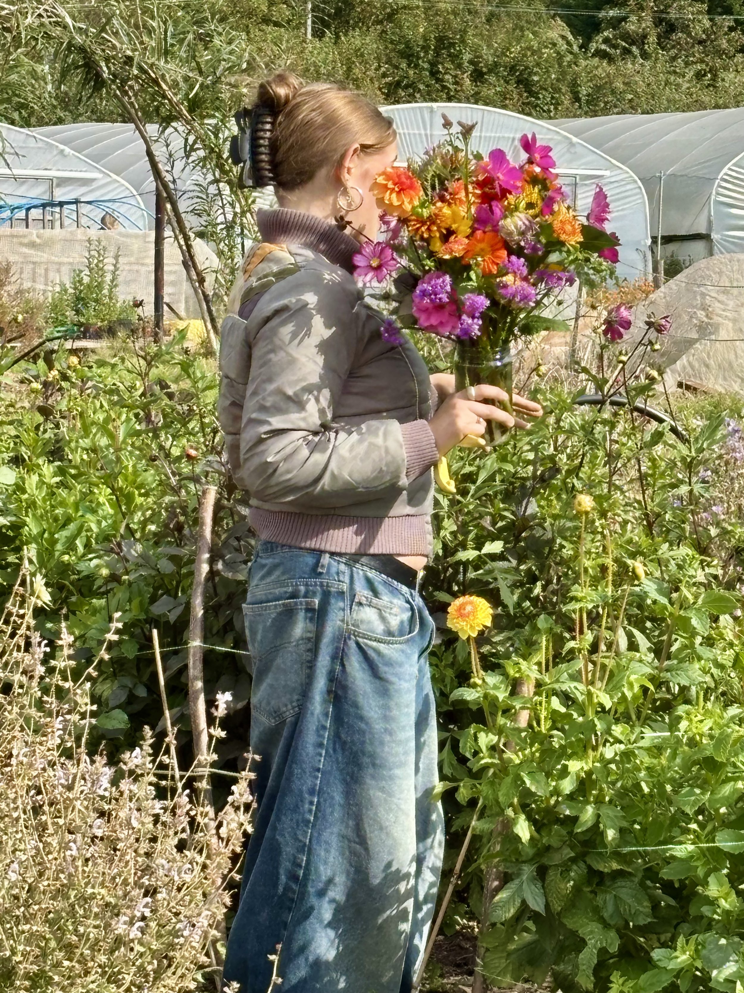 A woman with hair tied up, wearing a grey jacket with a pink sleeve and large hoop earring, holding a colourful bouquet of flowers at 5 Acre Farm with green plants and white greenhouses in the background.