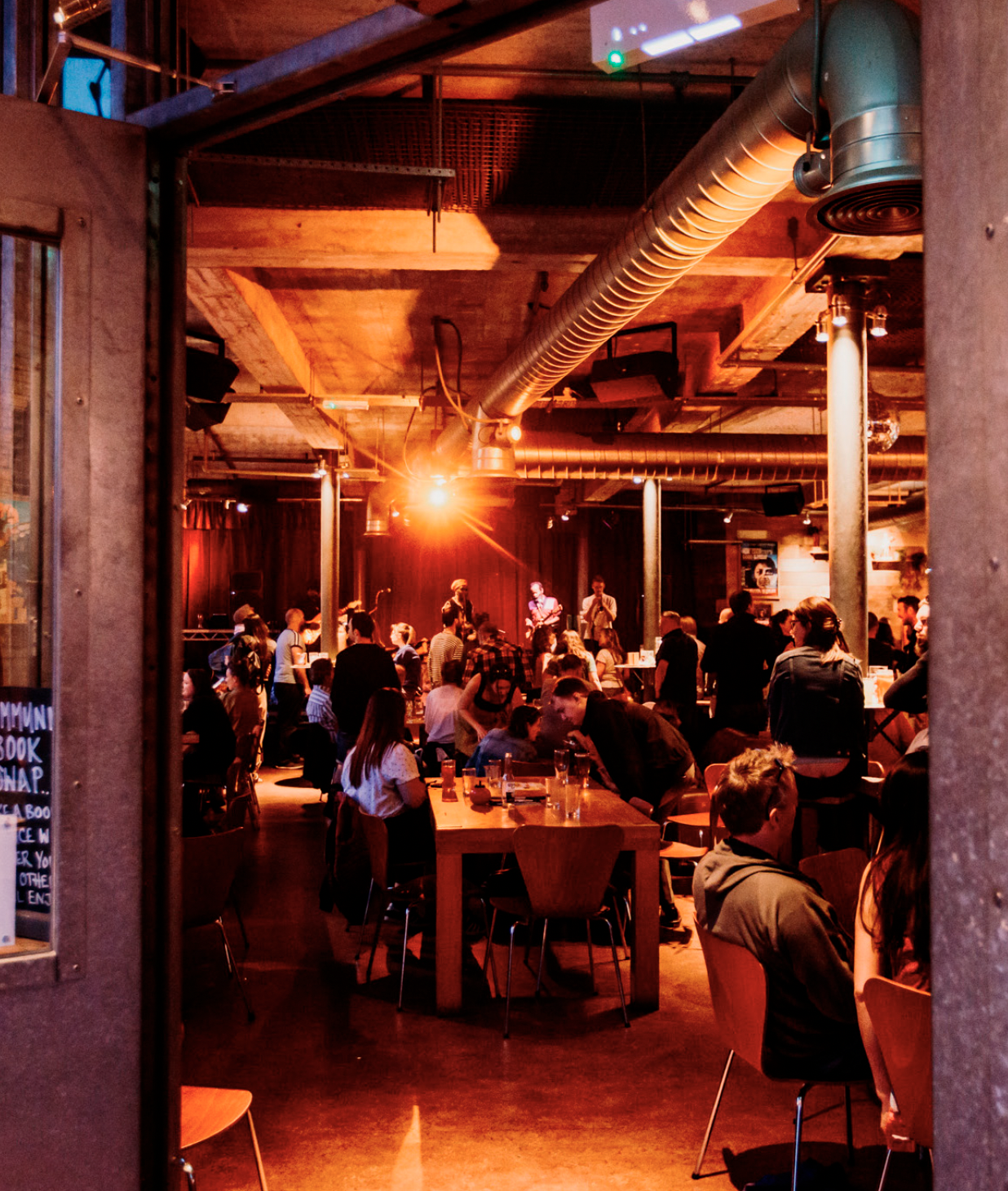 People gathered in the Cafe Bar to watch live music in the evening. There is a stage where a band is performing. The room features exposed wooden beams and large industrial ducts on the ceiling, with warm orange and red lighting creating a lively atm