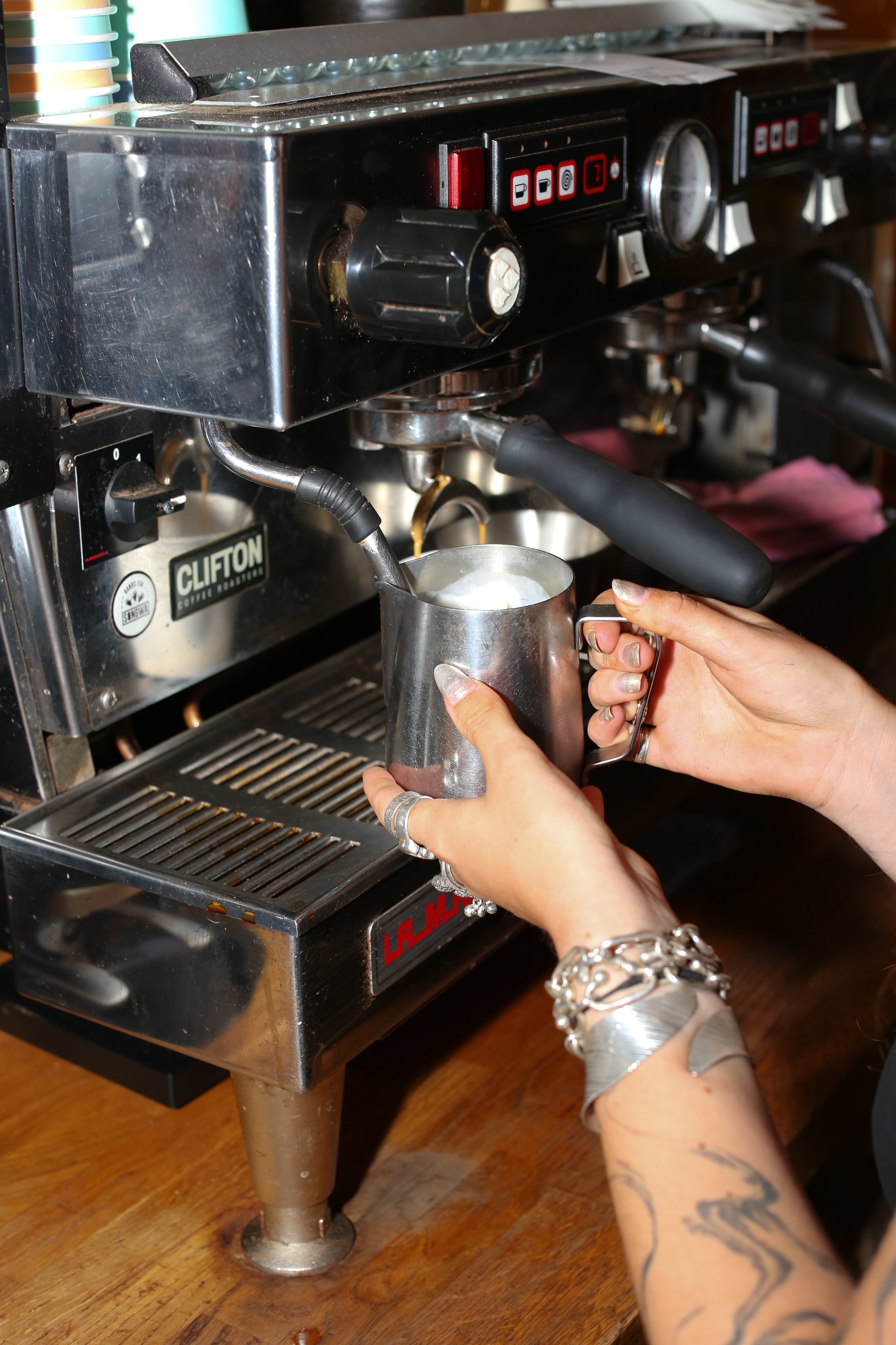 Barista making Coffee at Tobacco Factory. A person with tattoos on their arms and a bracelet is holding a metallic cup while operating a professional espresso machine to make coffee.