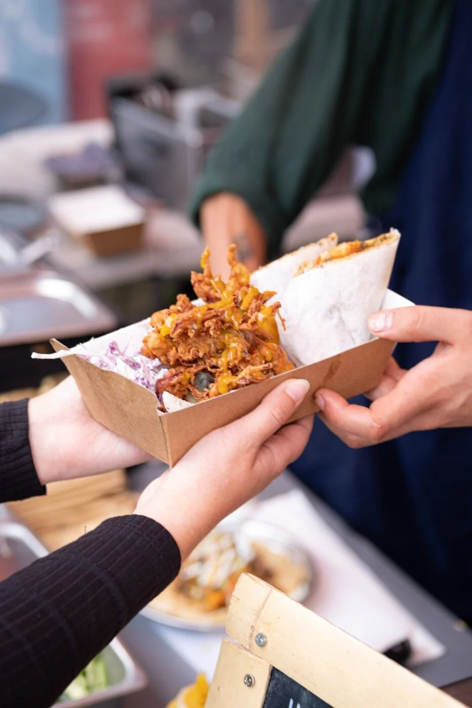 Person holding a cardboard serving tray with pulled pork sandwich and coleslaw in a food setup.