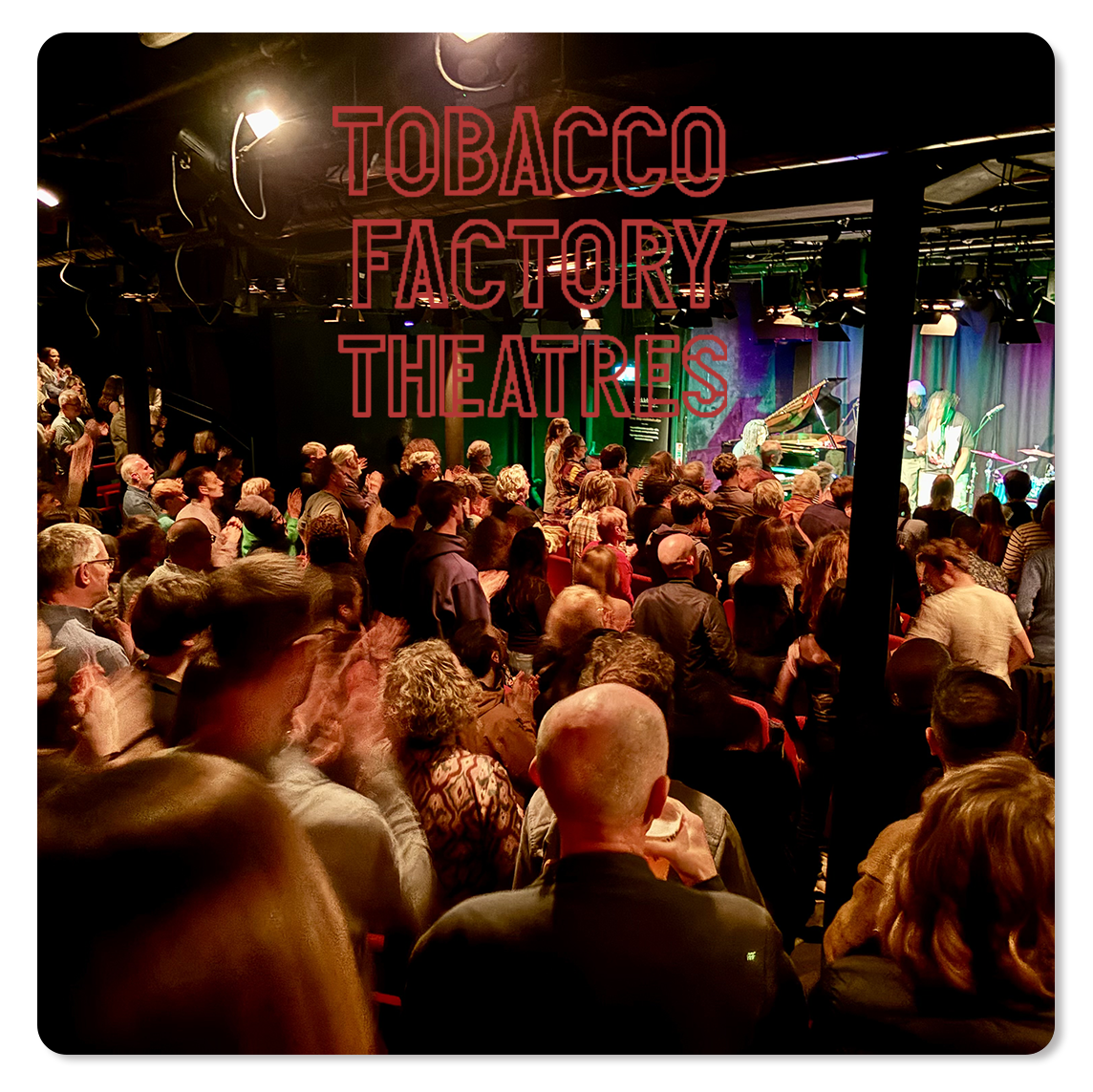Audience applauding at a concert with a band on stage, featuring a grand piano and microphones, at the Tobacco Factory Theatres.