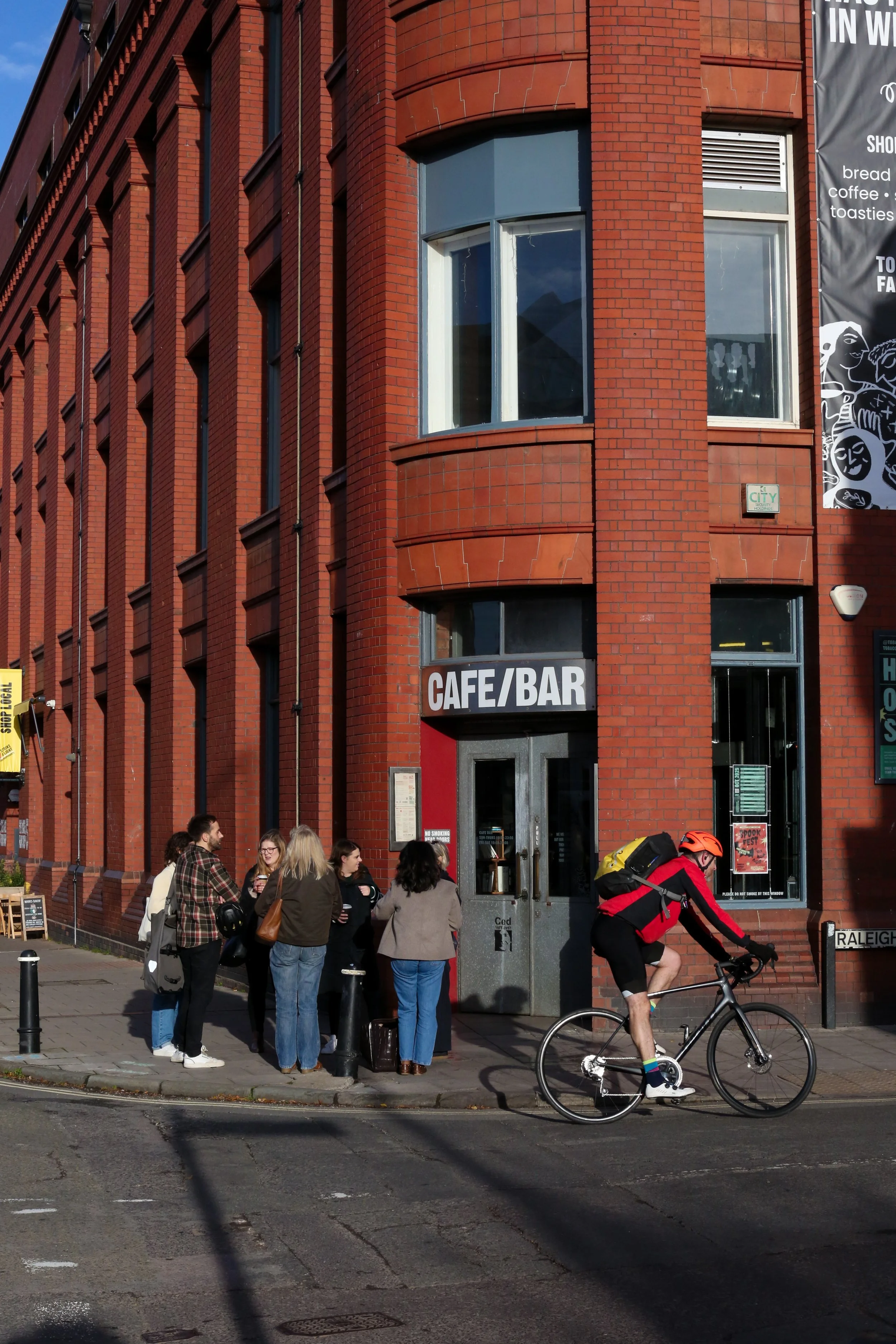 A group of people standing outside the CAFE BAR', while a cyclist rides by on the street. The building has large windows and a black door, and there are street signs and a banner on the wall.