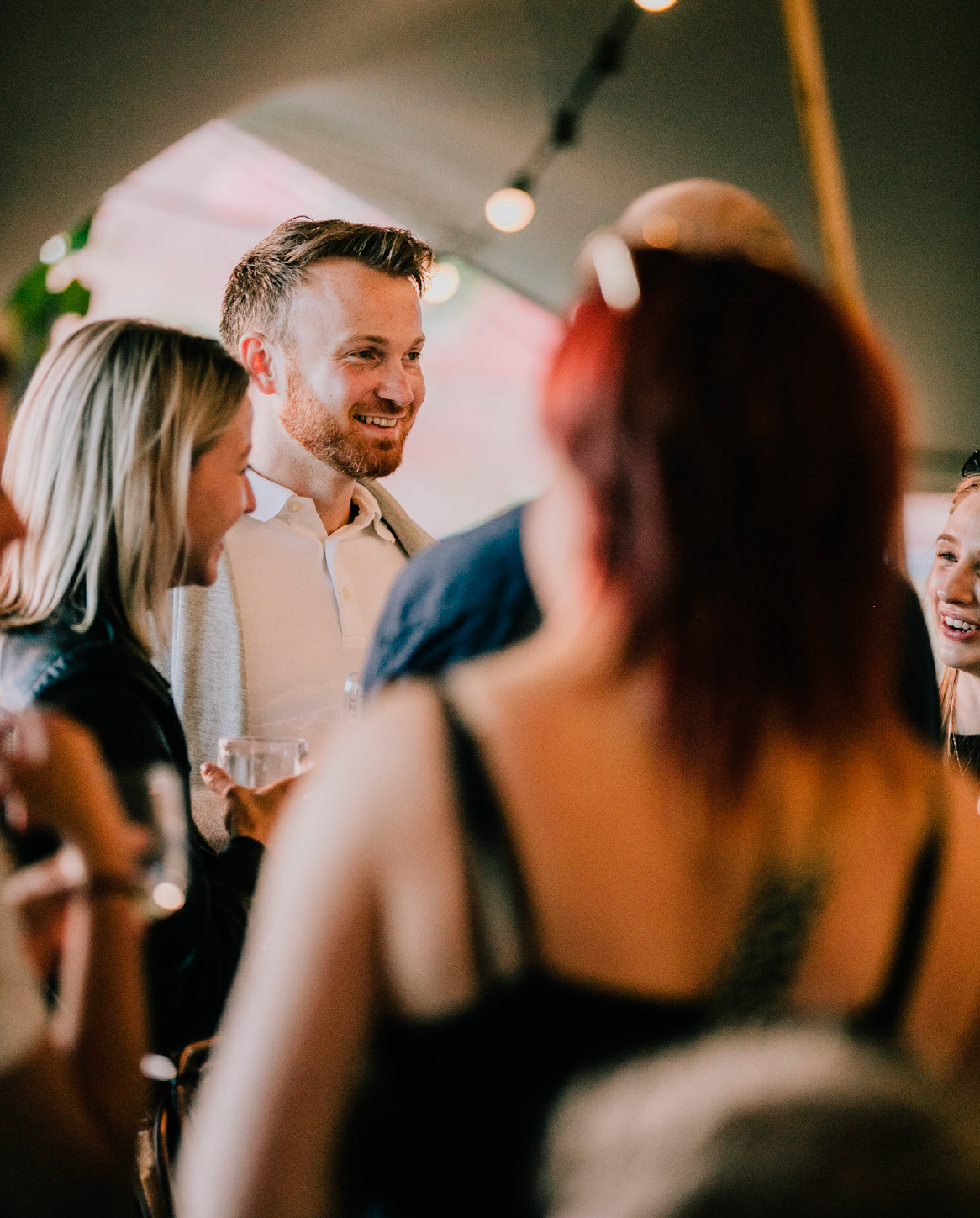 A group of people socializing indoors, smiling and holding drinks, with warm lighting and a lively atmosphere - hiring the tent & terrace for a private occasion.