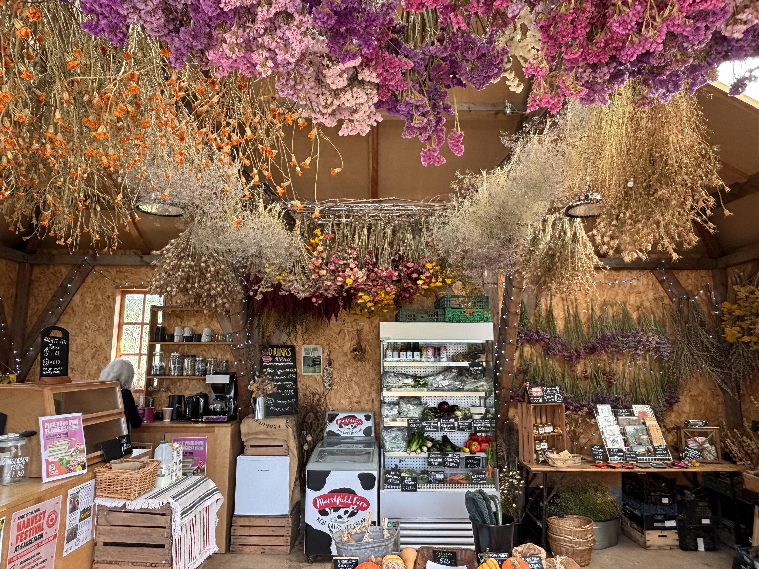 Indoor market stall at 5 Acre Farm with hanging dried flowers and herbs on the ceiling, shelves with produce, jars, and handwritten signs.