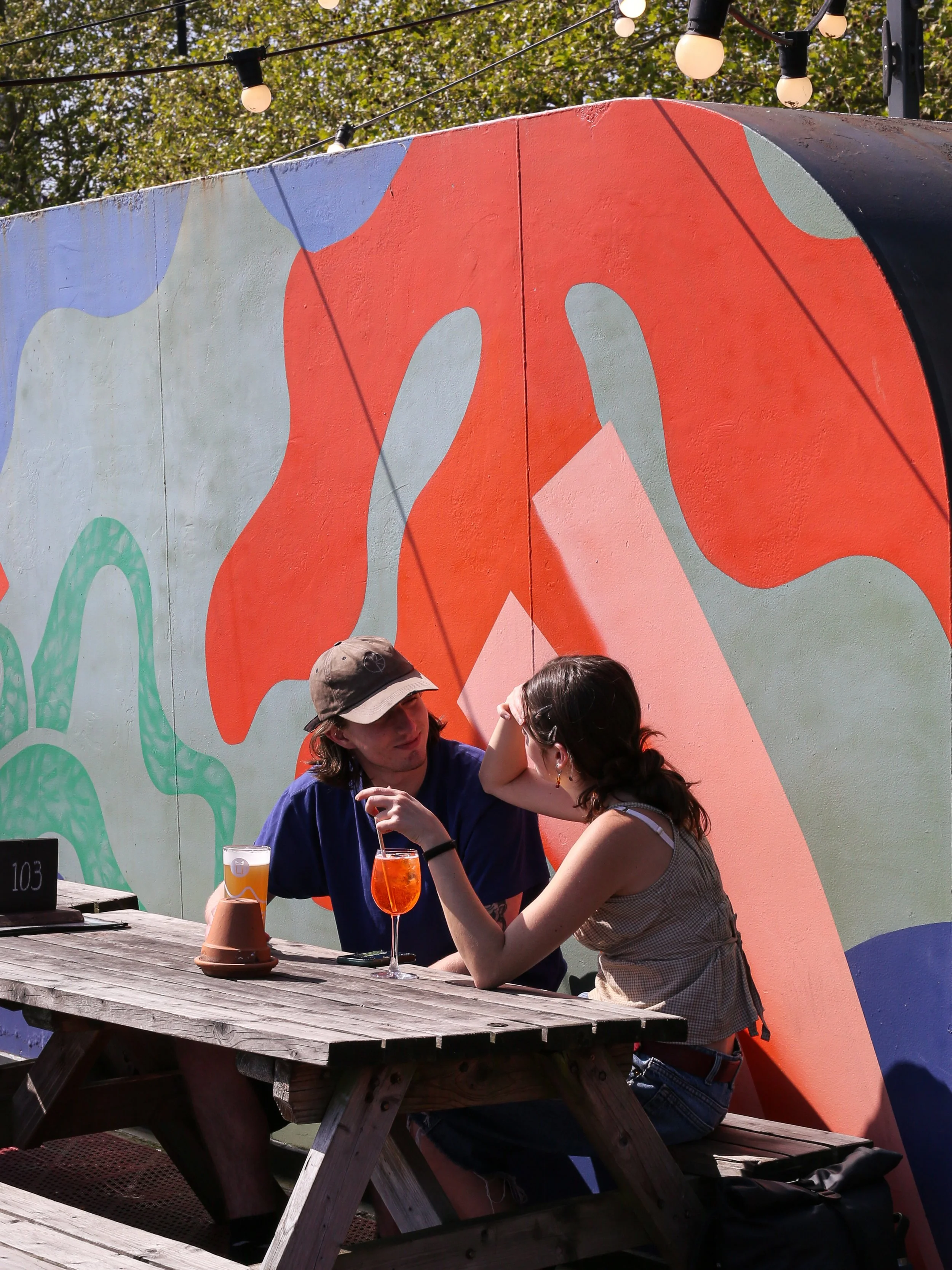 A man and a woman sitting at a wooden picnic table outdoors at Grain Barge in sunlight, engaged in conversation. They each have a drink in front of them and are leaning towards each other. Behind them is a colourful mural wall with abstract shapes an