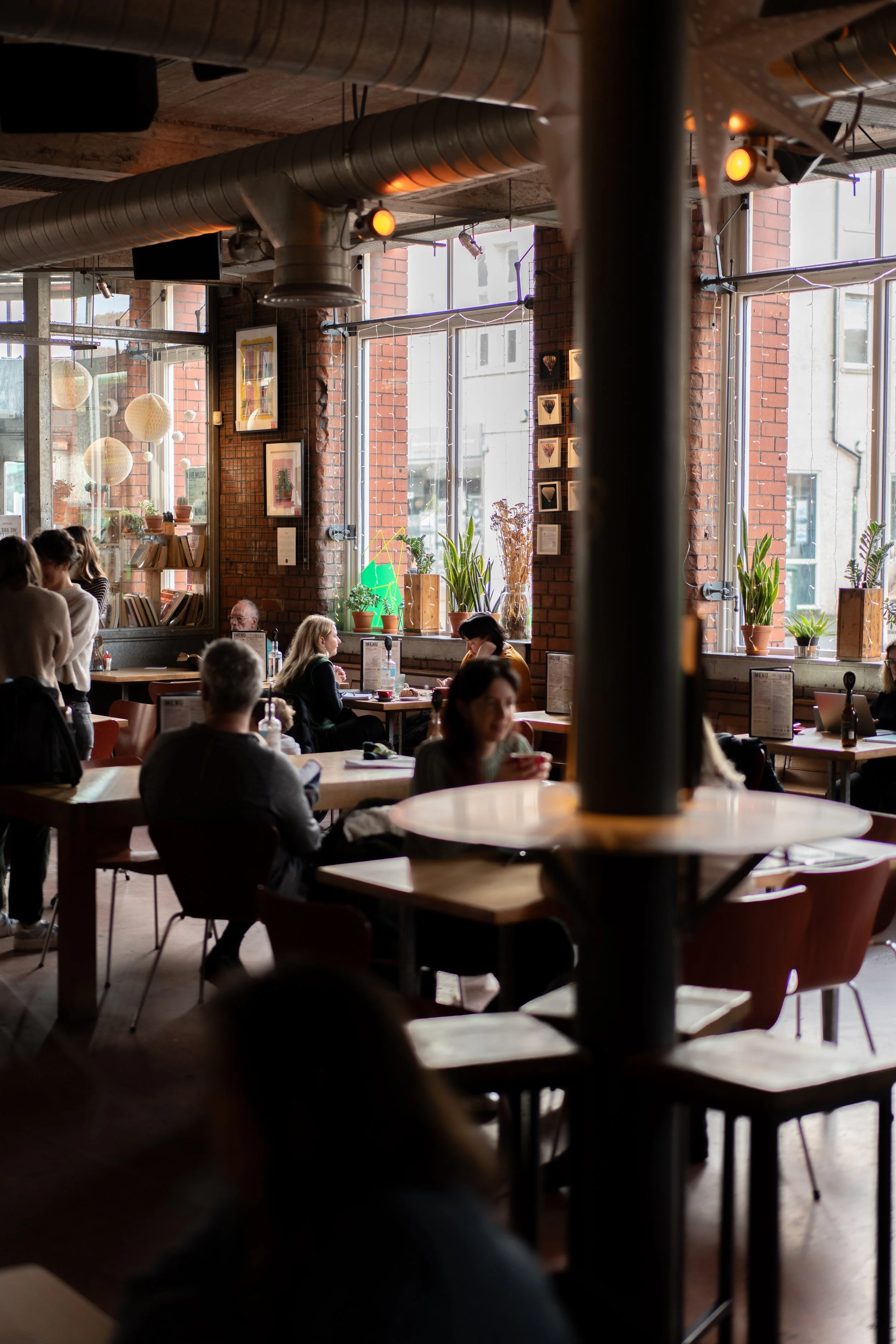 Interior of the cozy cafe bar and restaurant with large windows, brick walls, potted plants, framed pictures on the wall, and people sitting at tables enjoying their meals or drinks.
