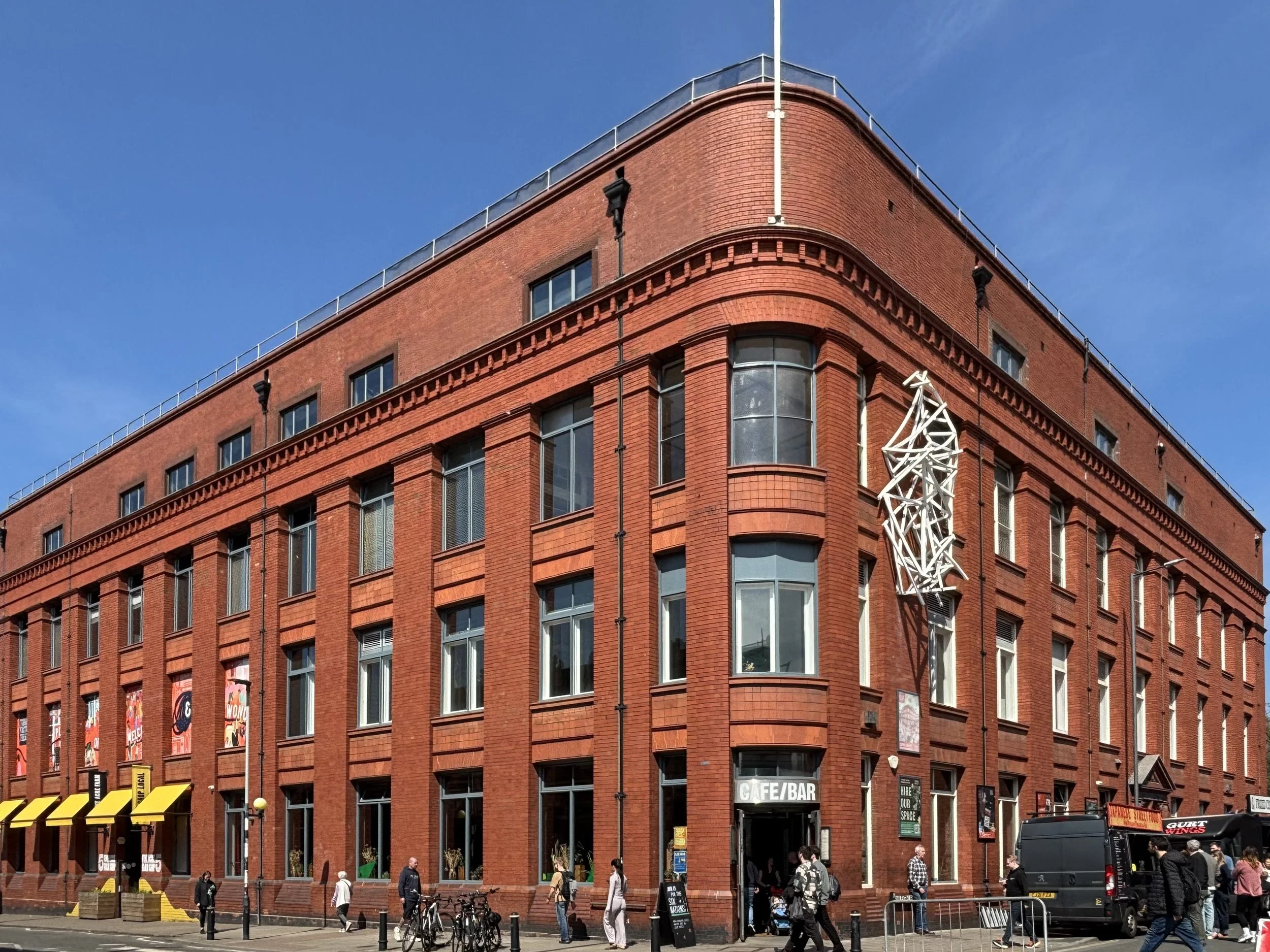 Red brick building with multiple windows and a Exterior shot of the corner of the road where North Street and Raleigh Road meet. The curved corner is the entrance to the Cafe Bar, with people walking on the pavement and bicycles parked in front, agai