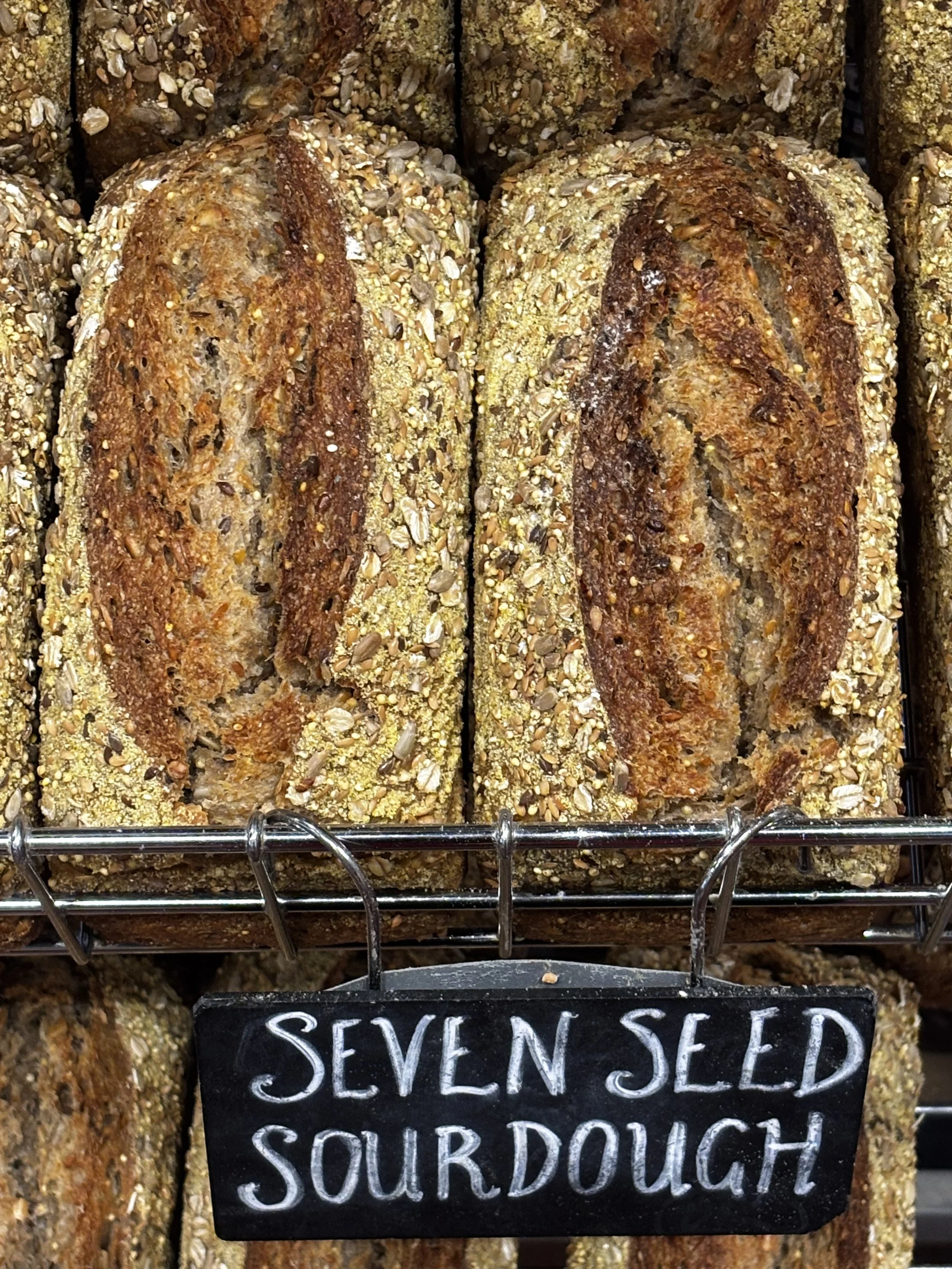 Close-up of bread from Mark's Bakery with a seed and grain crust, displayed behind a sign that reads "Seven Seed Sourdough".