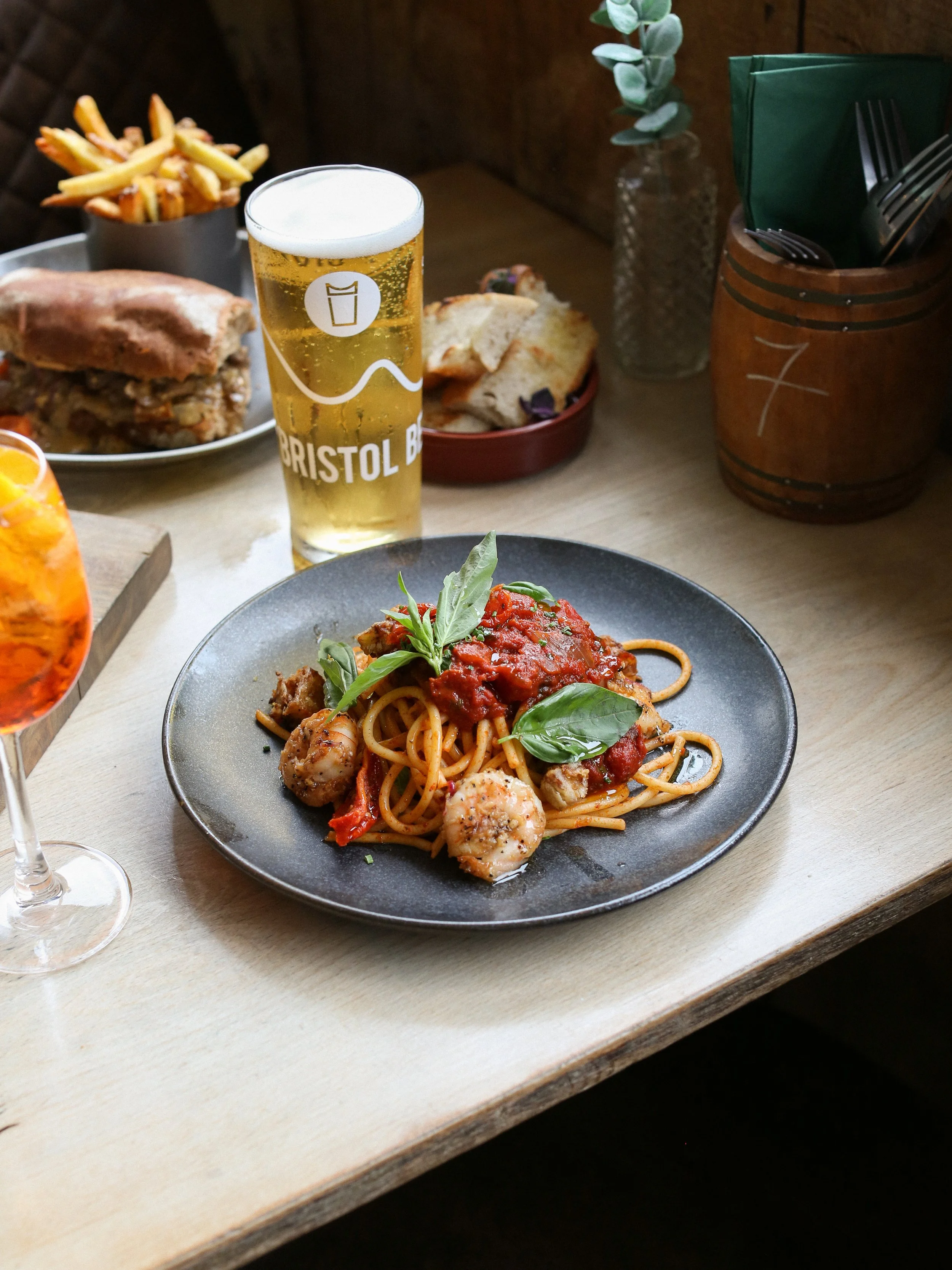 A plate of spaghetti with prawns and fresh basil, a glass of beer, fries, a burger, and a bowl of bread on a table at a restaurant.
