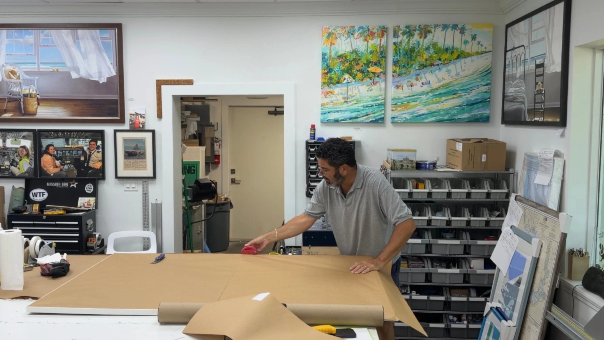 Man standing at a work table wrapping large sheet of brown paper, surrounded by art supplies and framed pictures on the wall. 	
Sanibel Framing. Custom Frames.  Paul Roshburg
